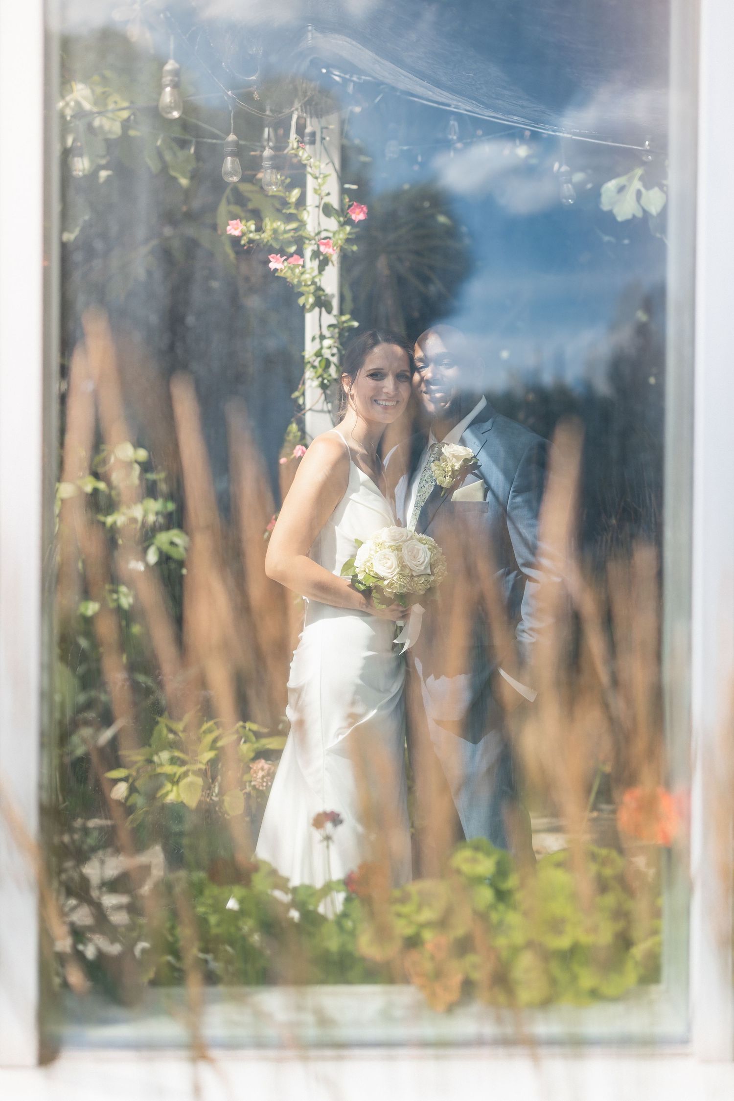 Artistic reflection of wedding couple through greenhouse windows with soft natural lighting.
