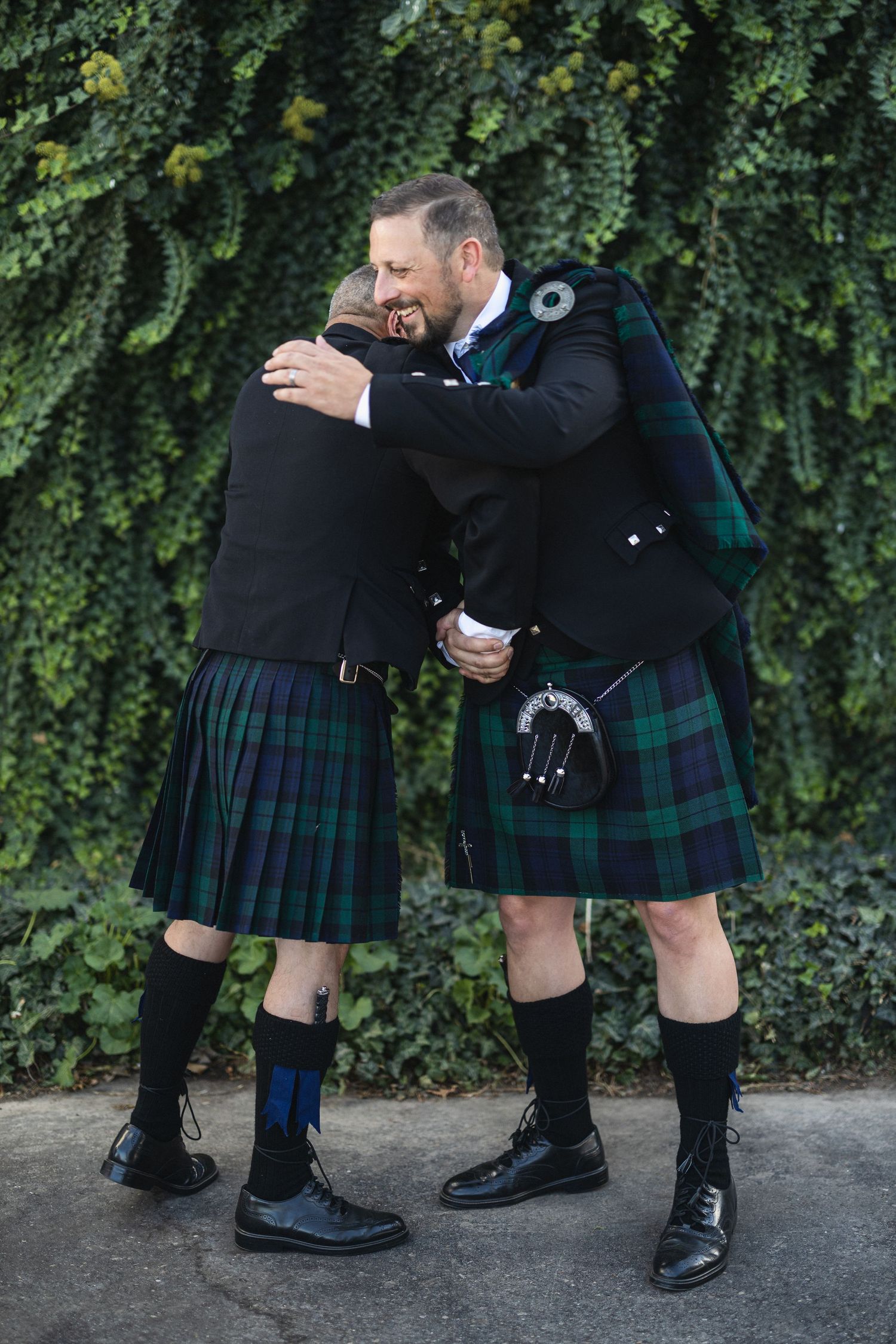 Two men in traditional Scottish kilts embrace warmly against ivy wall.