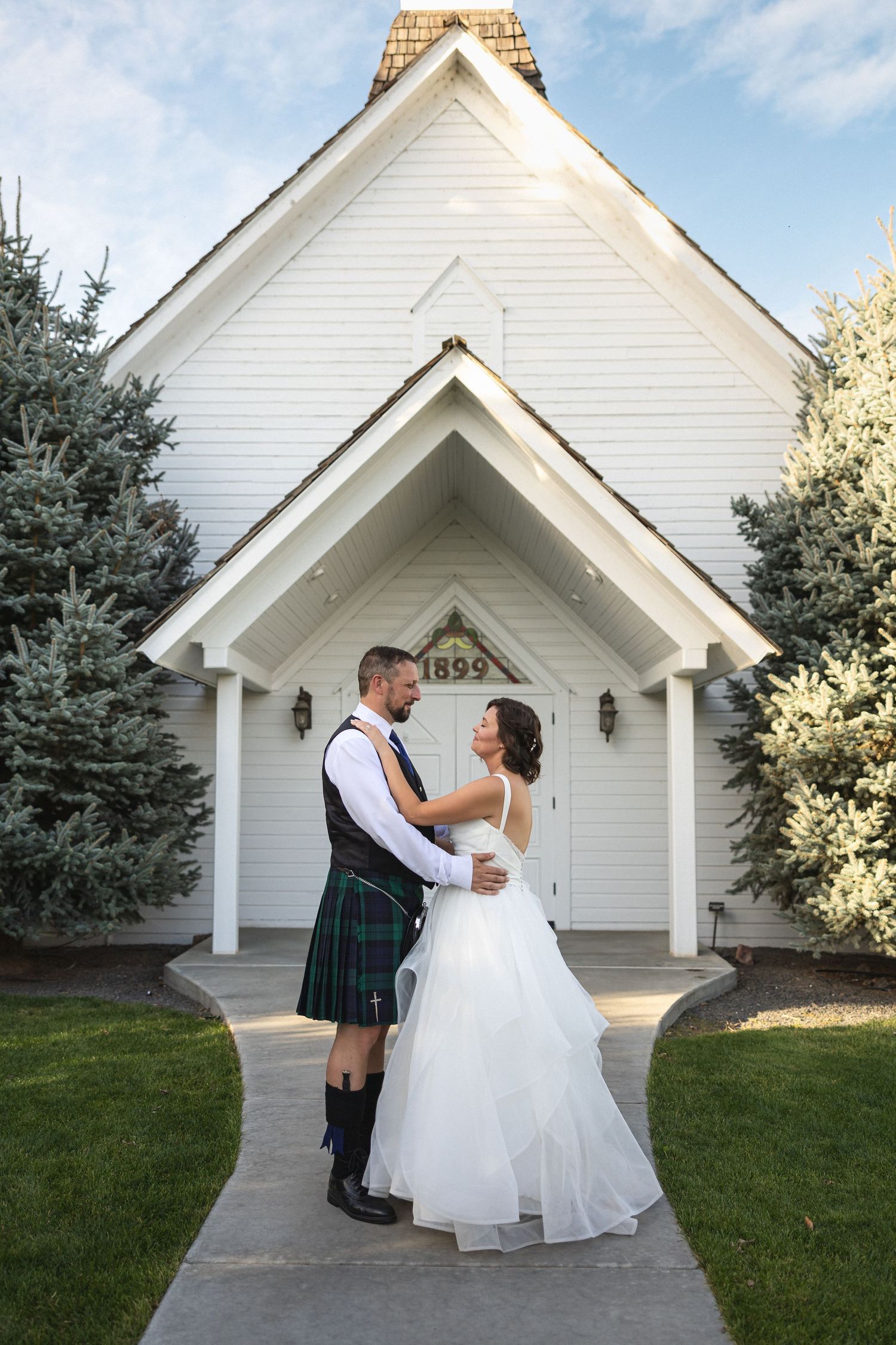 Couple in Scottish wedding attire embraces in front of white chapel entrance.