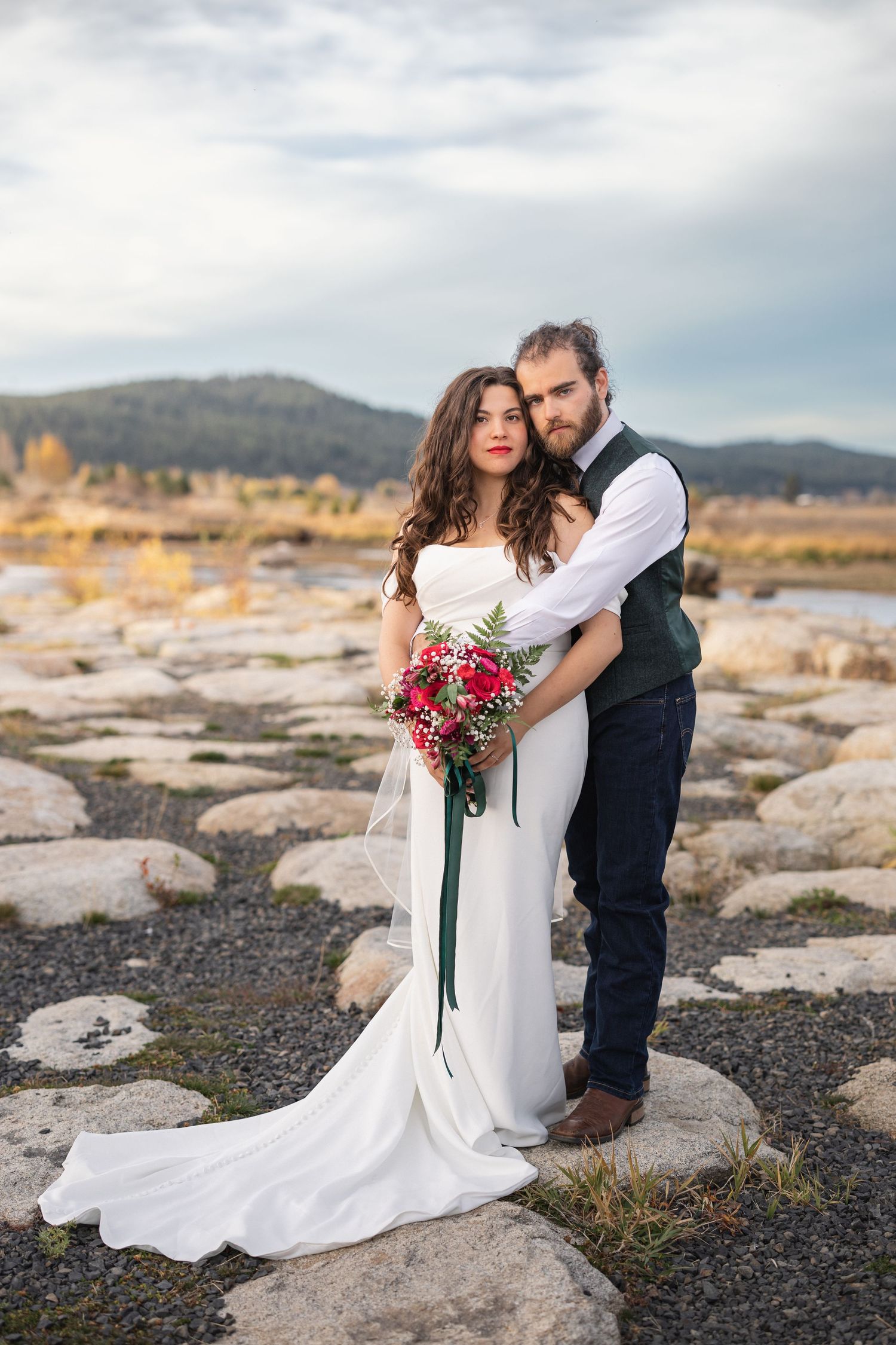 Wedding couple poses against mountain landscape with red bouquet and flowing white dress.