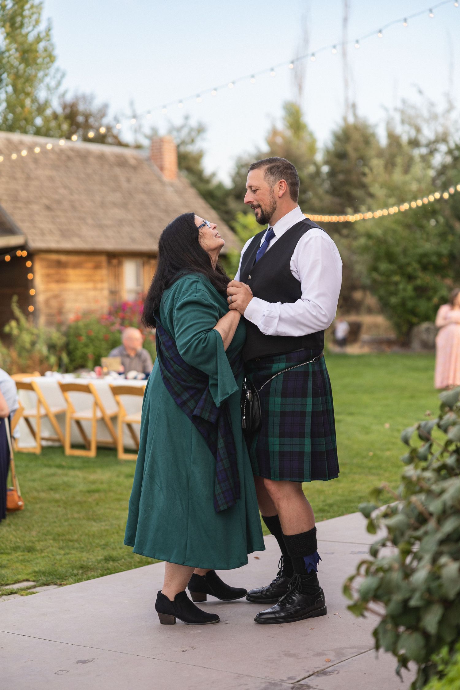 A couple shares a tender moment outdoors with string lights and rustic buildings in the background.