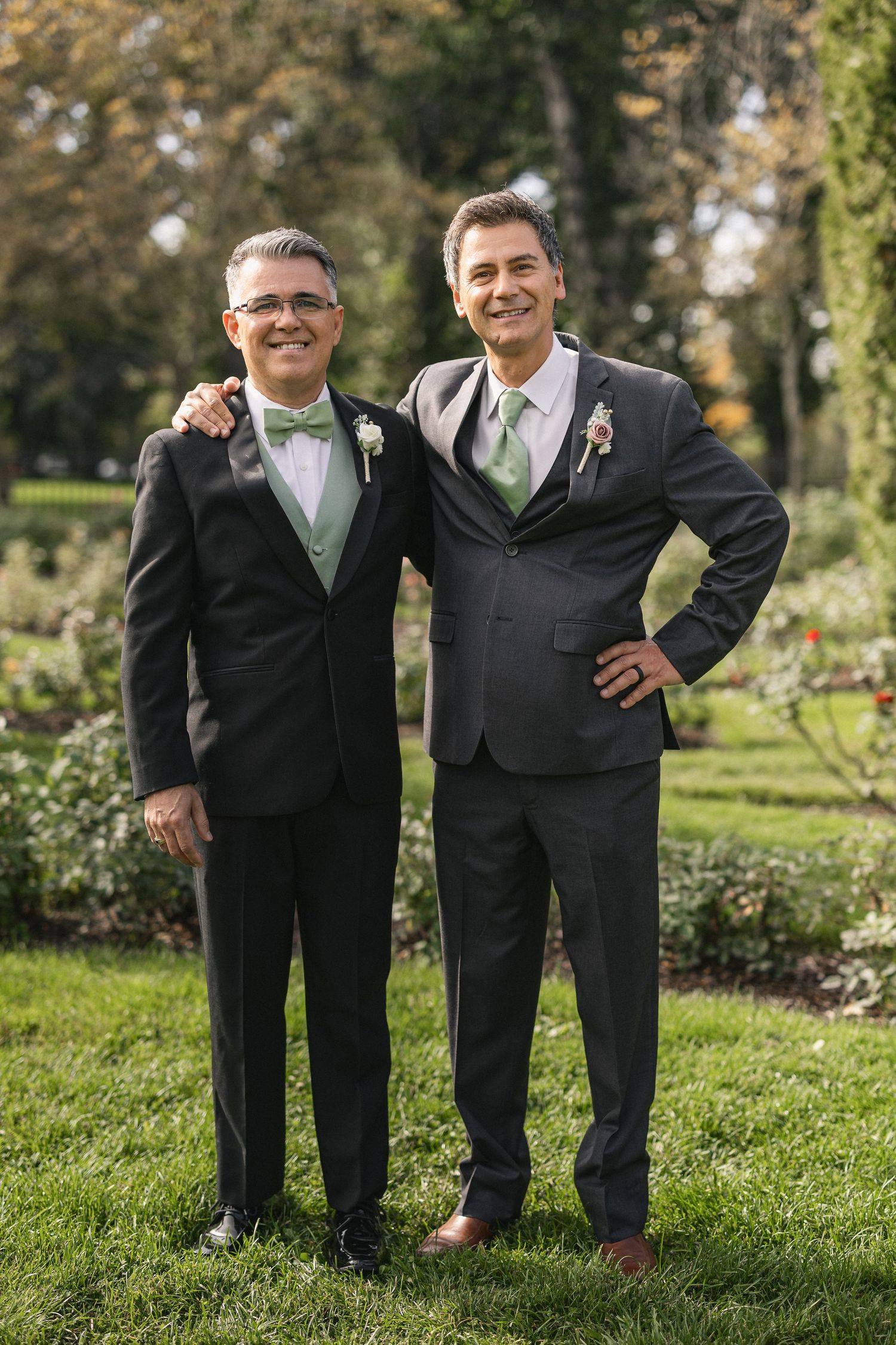 Two men in matching dark suits and green accessories pose together at a garden wedding.