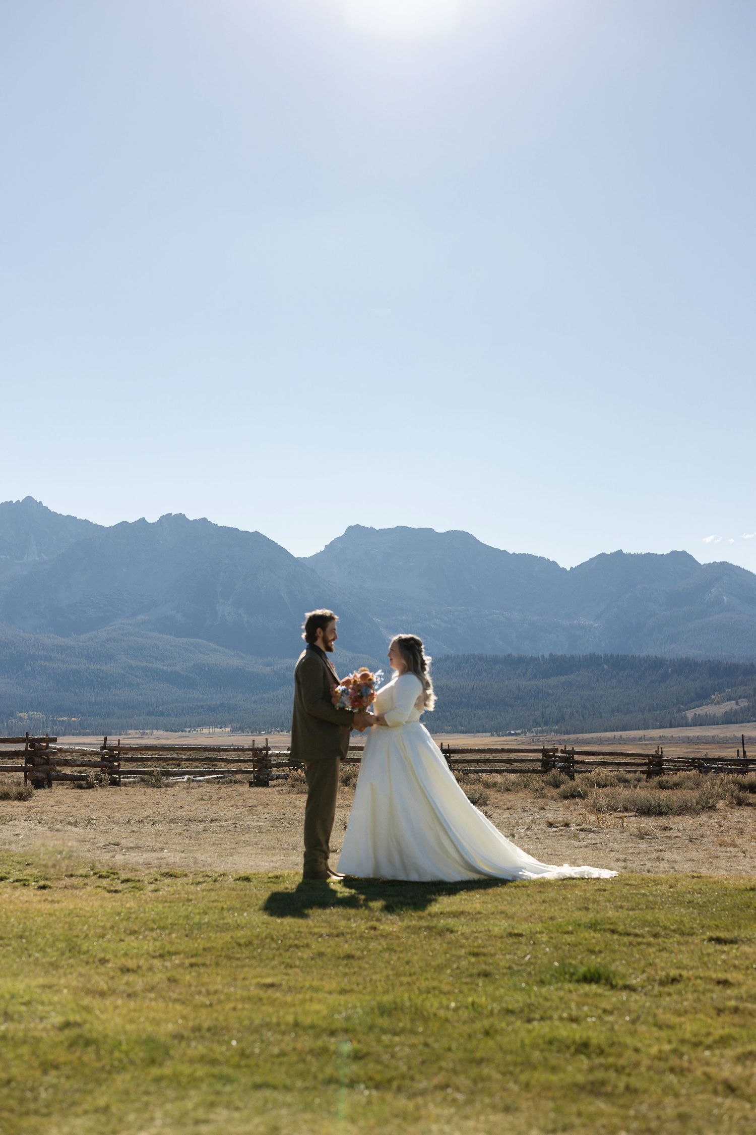 A wedding couple stands in an open field with majestic mountain peaks rising in the distance.