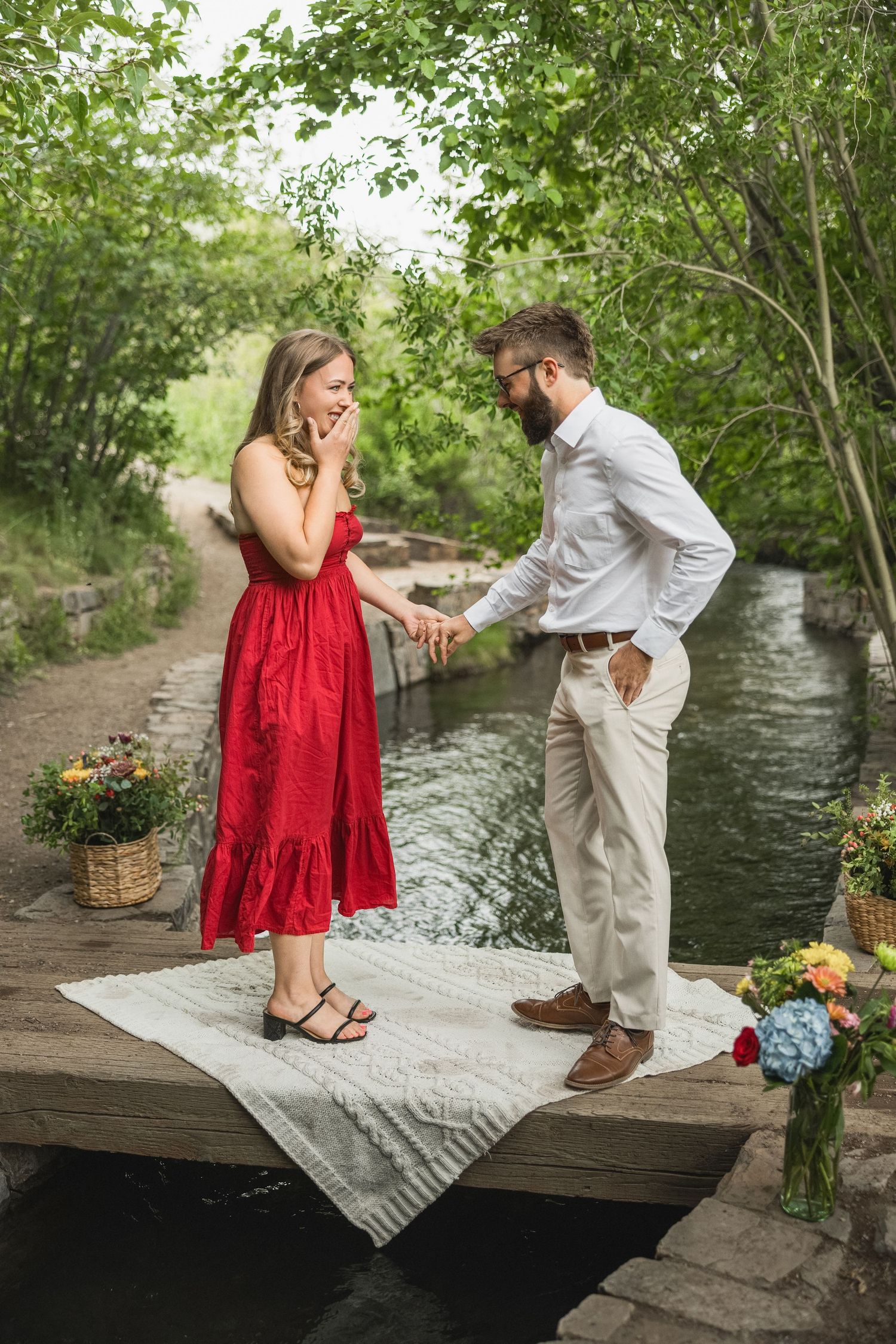 A romantic proposal moment on a bridge over a stream, with a person in red dress reacting to their partner kneeling.