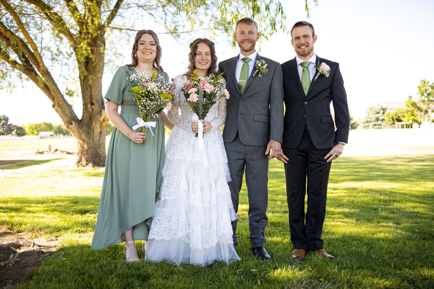 Small wedding party poses under tree with bride in white gown and attendant in sage dress.