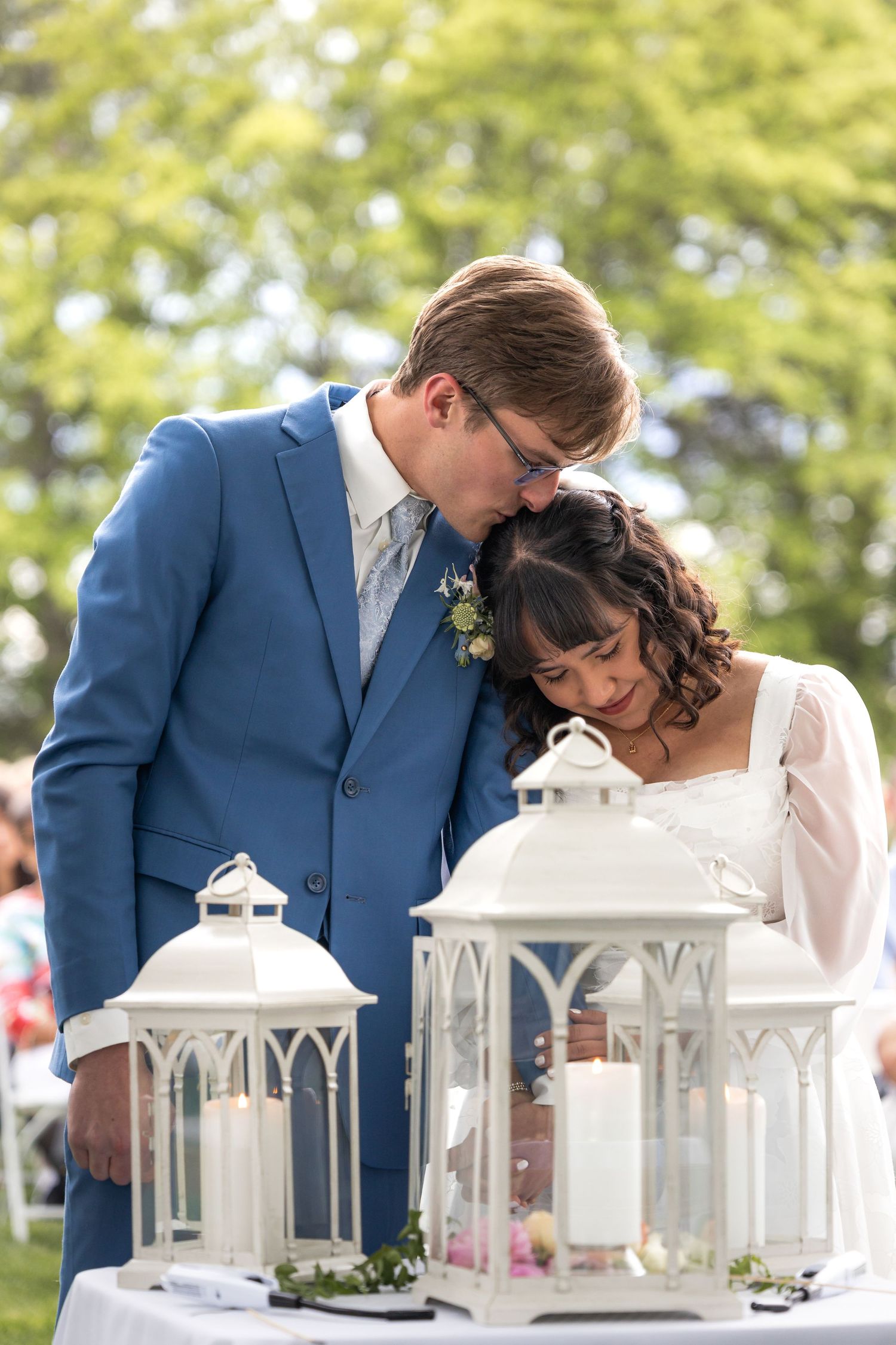 Tender moment between couple in blue suit and white dress with lantern decorations at outdoor wedding.