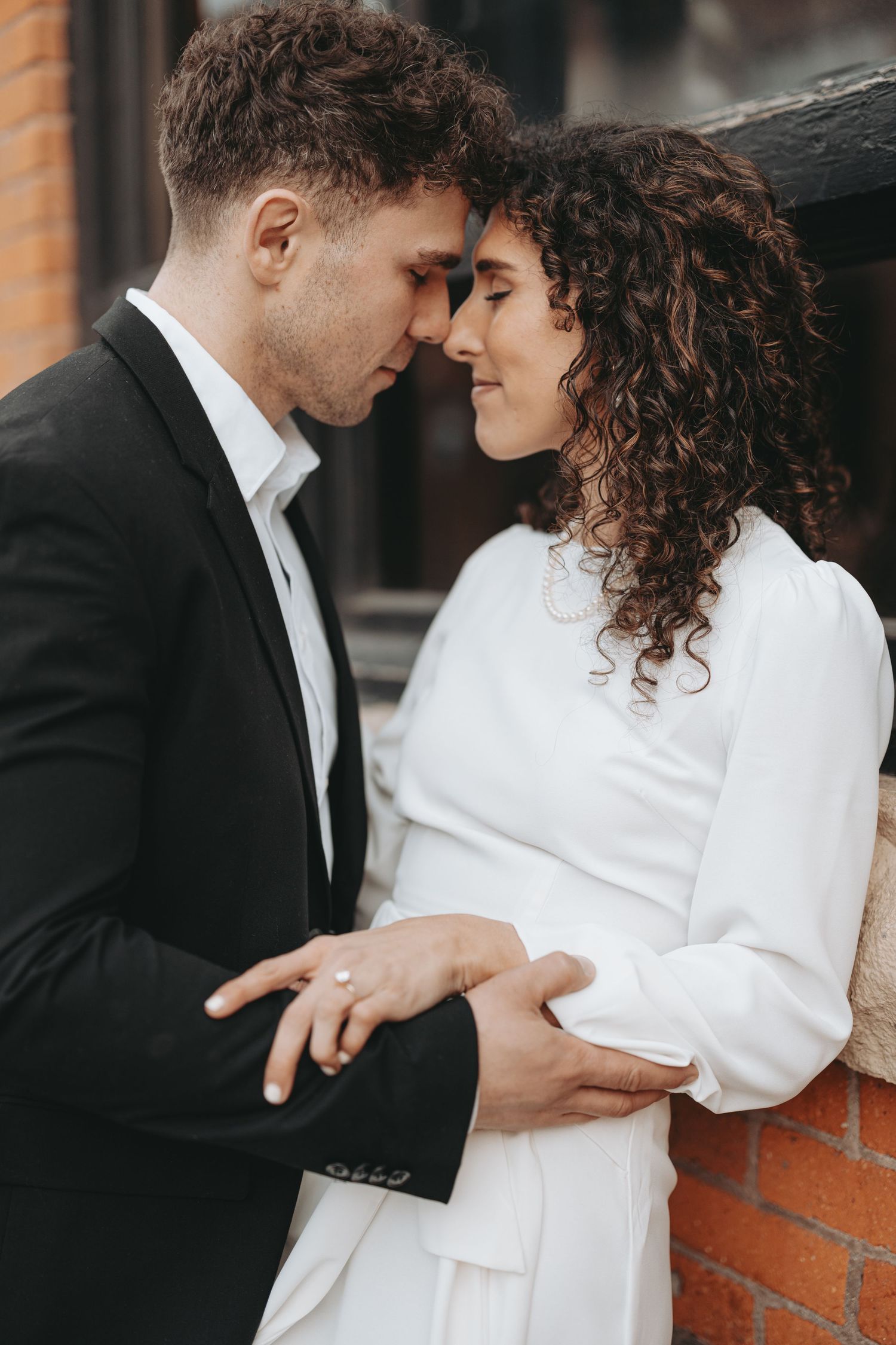 An intimate moment between a couple in formal attire against a rustic brick backdrop.