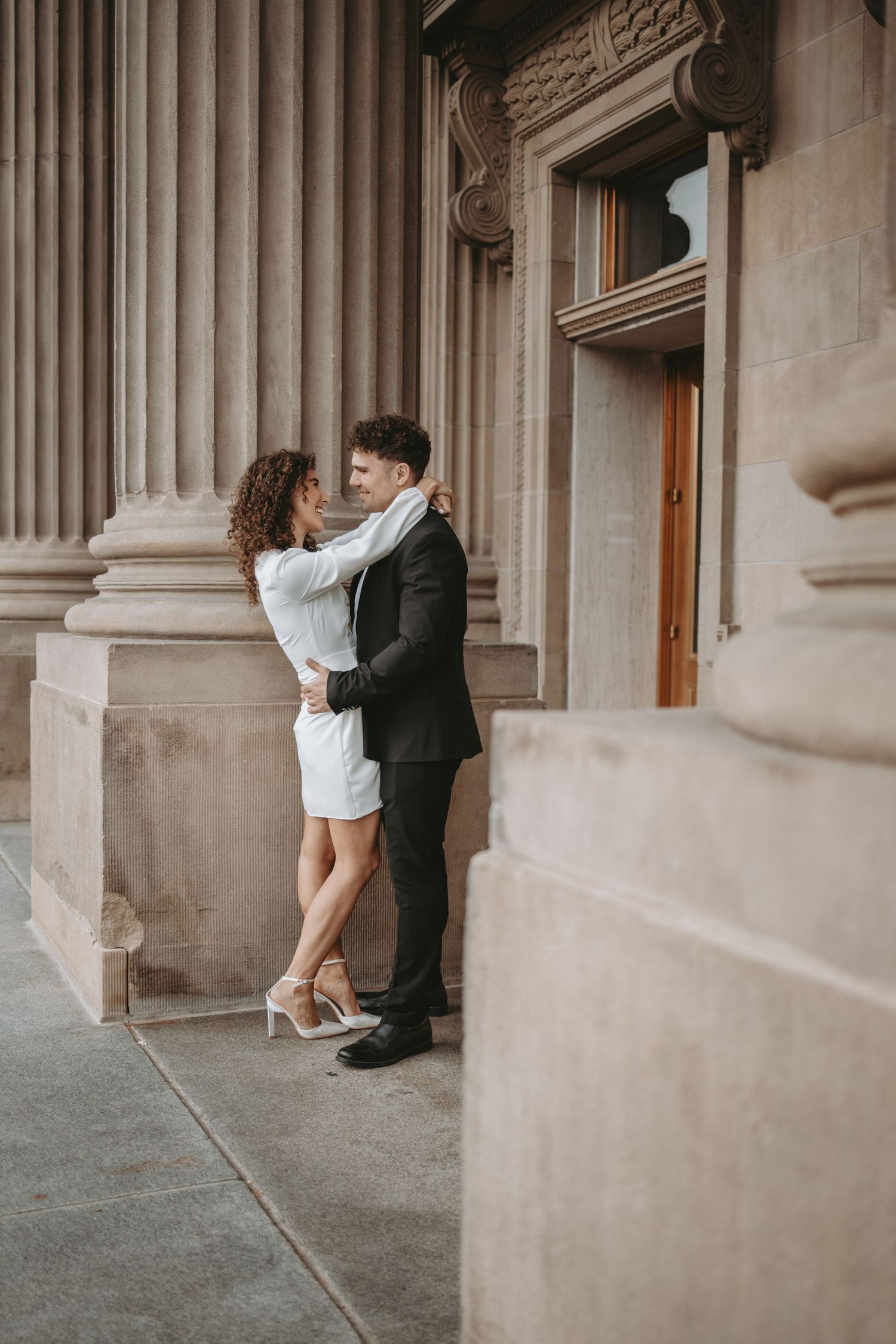 A couple in formal attire shares an intimate moment between classic stone columns.