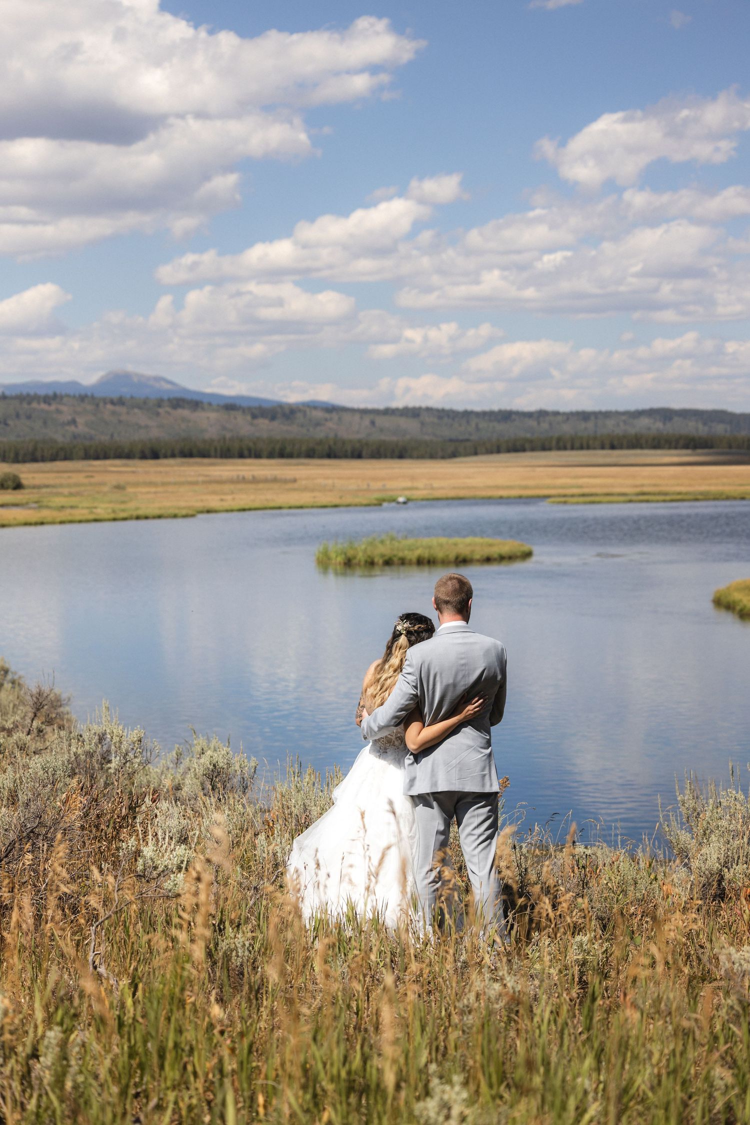Newlyweds share an intimate moment by a tranquil lake surrounded by tall grass and mountains.