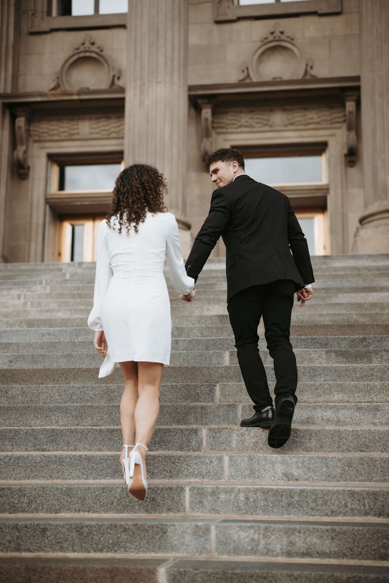 A couple in formal attire climb stone steps together outside a grand building with ornate architecture.