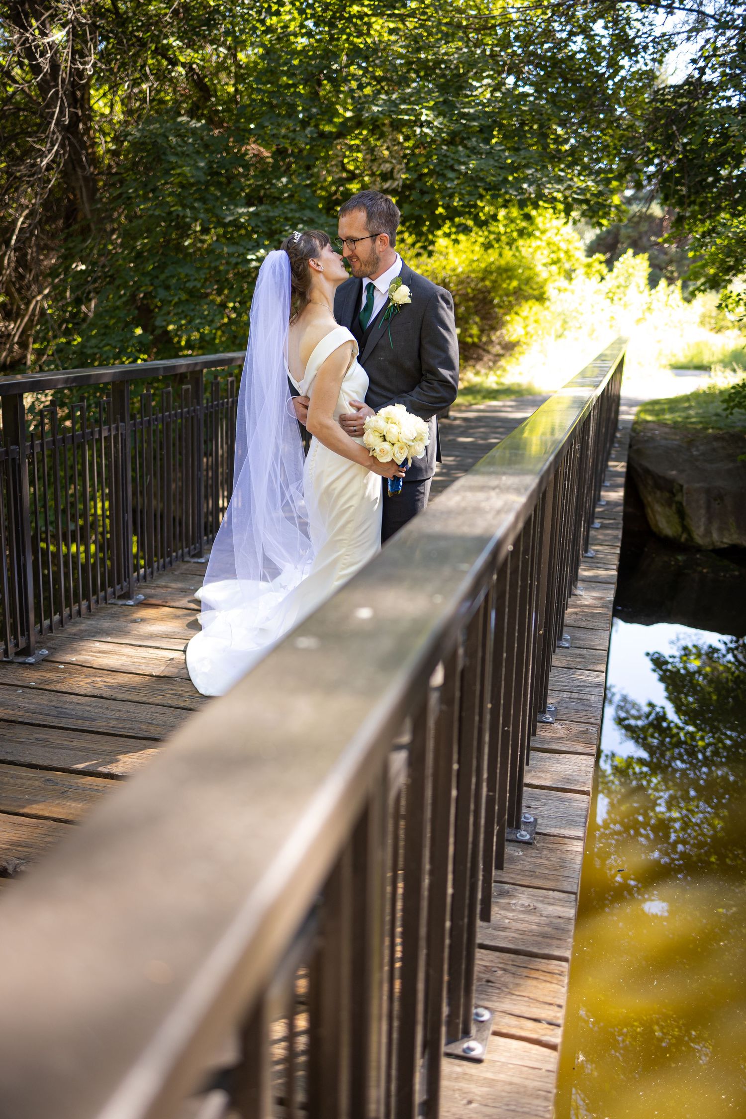 Wedding couple poses on wooden bridge with flowing veil and natural backdrop.