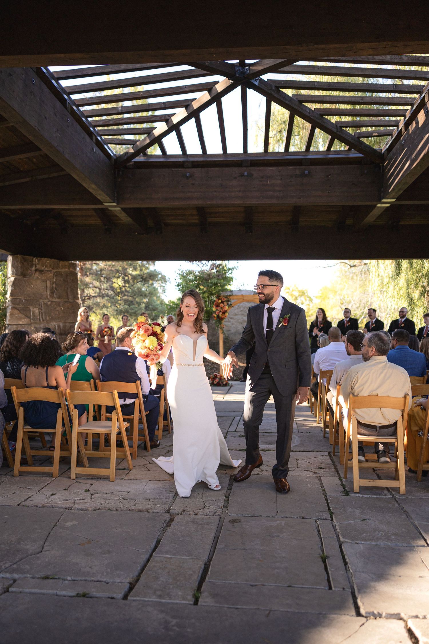Wedding couple walks down stone aisle beneath rustic wooden pergola during outdoor ceremony.