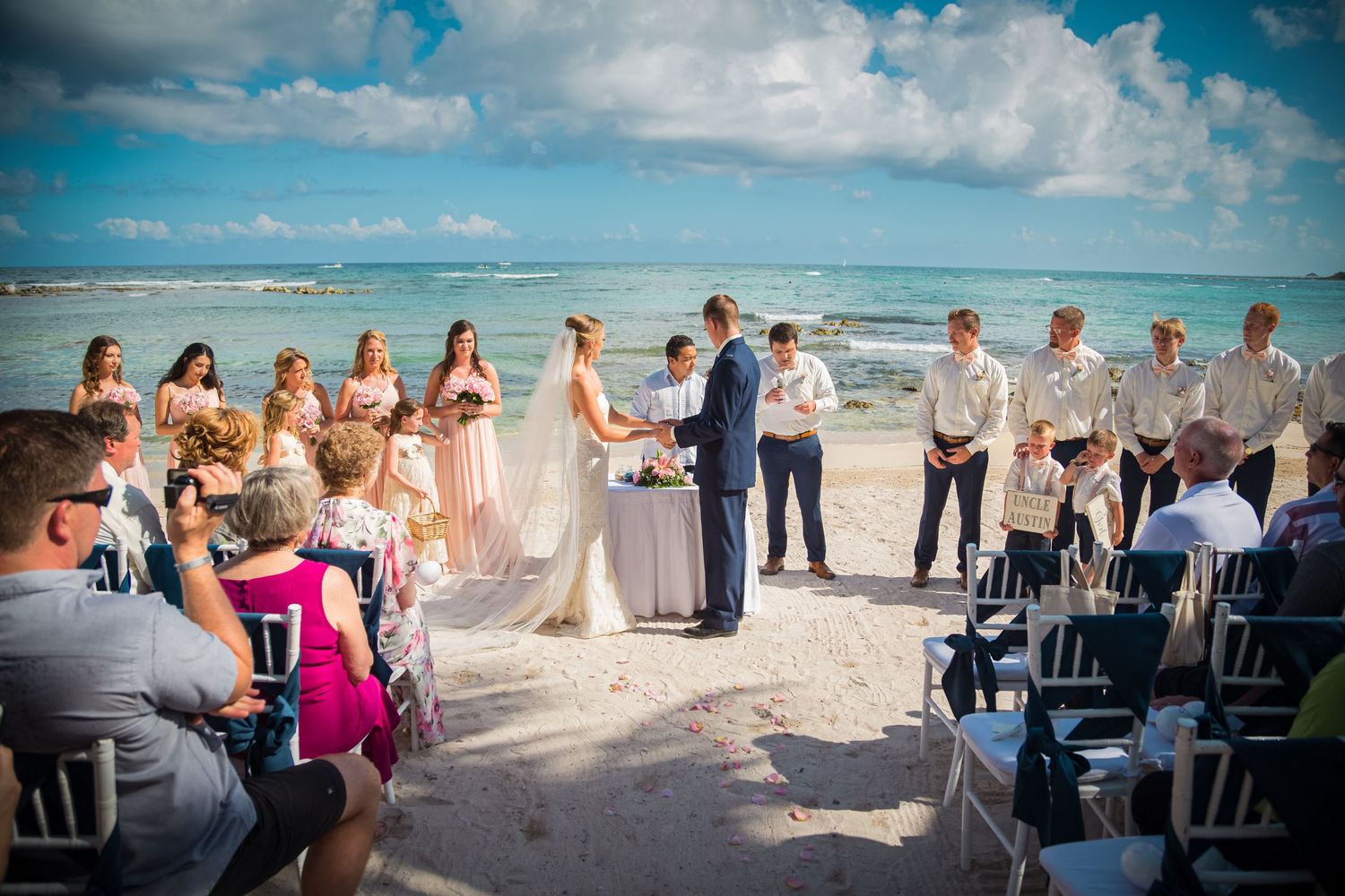 A beachside wedding ceremony takes place on white sand with turquoise ocean waves and blue sky in the background.