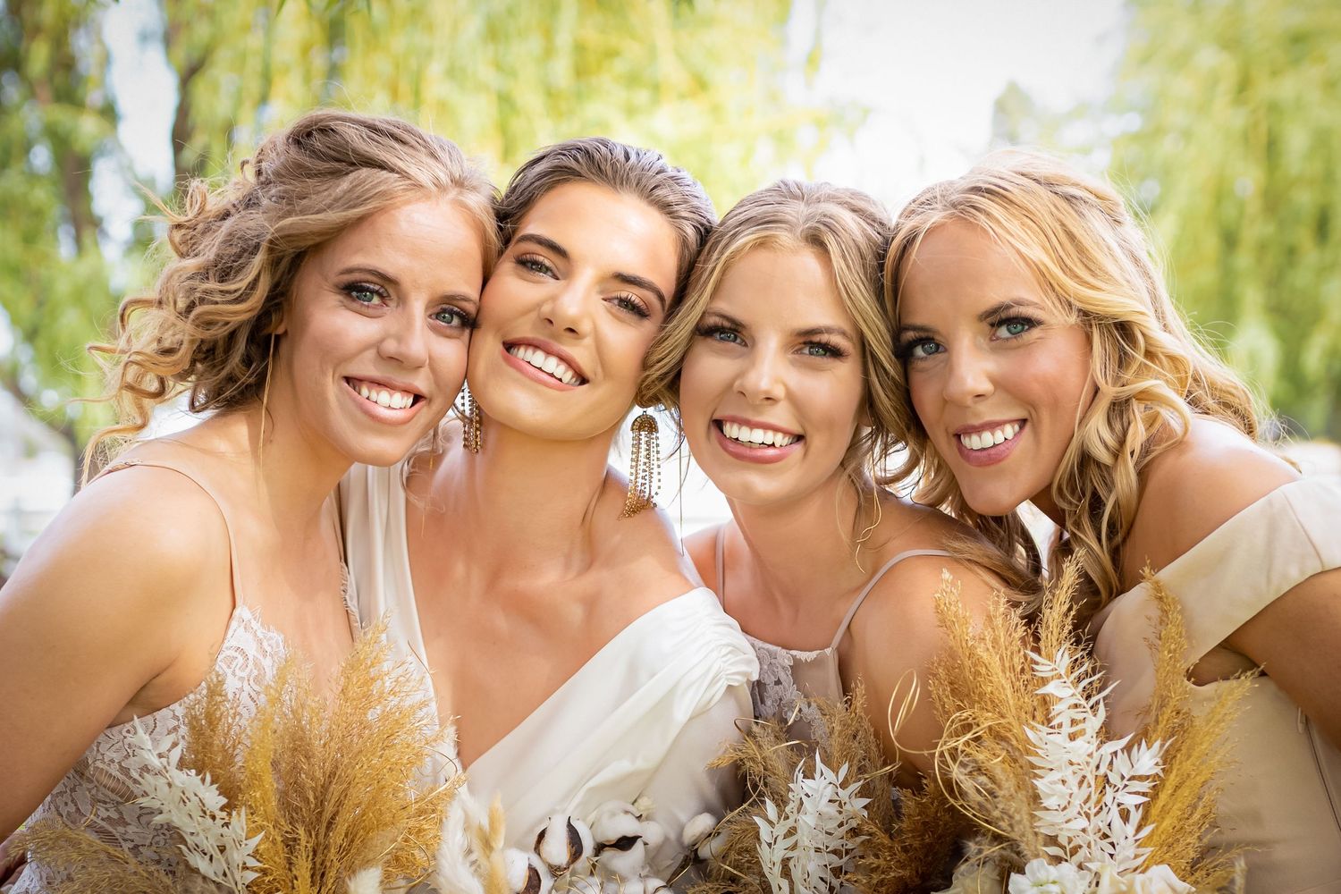 Bridesmaids with floral bouquets huddle close together under willow tree for joyful photo.