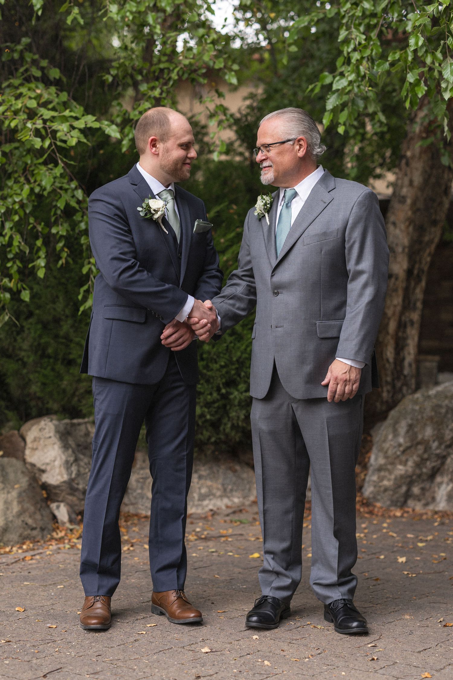Two men in suits share handshake during wedding celebration outdoors.