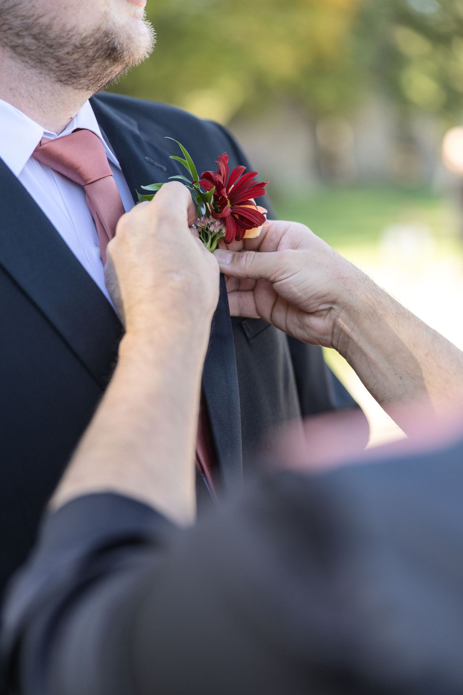 Hands pinning burgundy and peach floral boutonnière to suit lapel.
