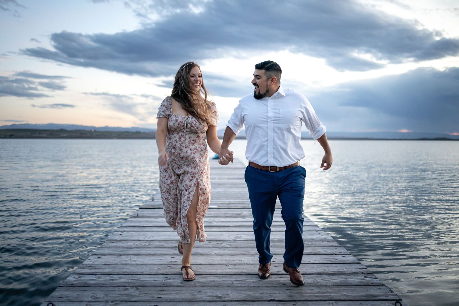 A couple walks hand in hand along a wooden pier with a dramatic cloudy sunset over the water.