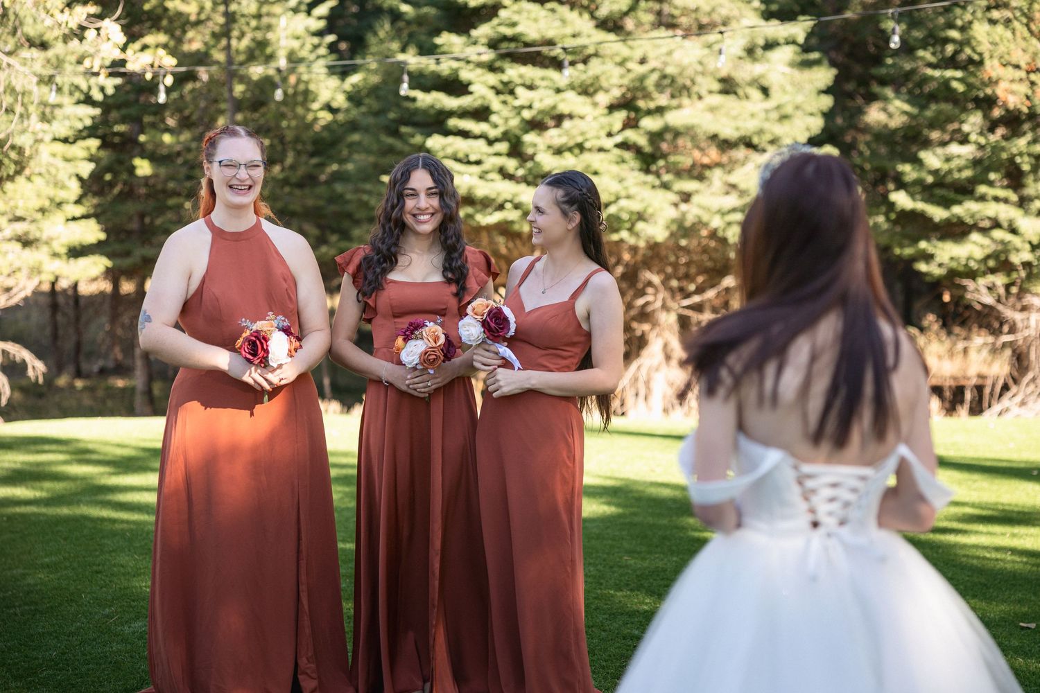 Bridesmaids in rust-colored dresses watch bride in white dress during outdoor photo session.