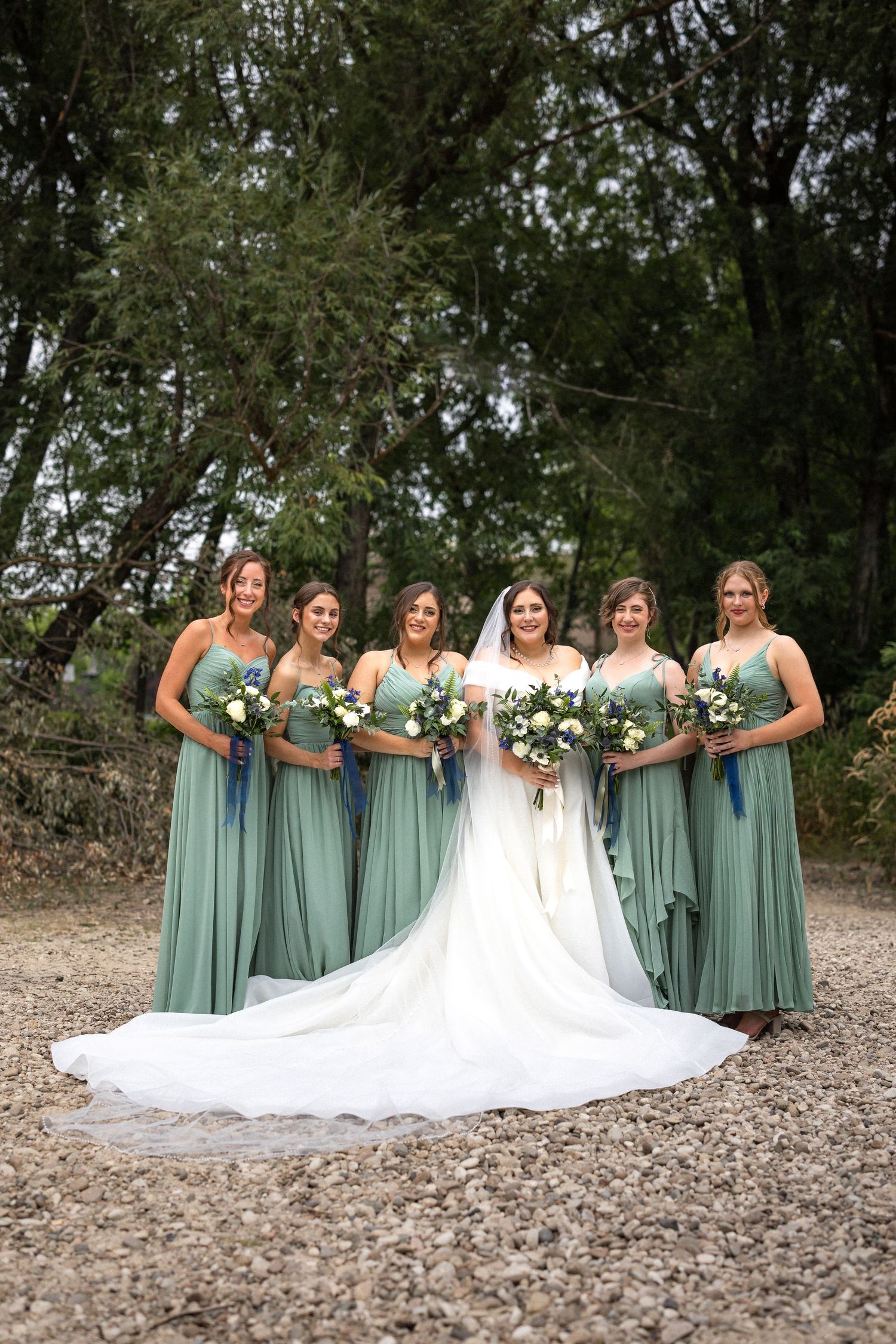 Bridesmaids in sage green dresses pose with bride wearing a long train white wedding gown.