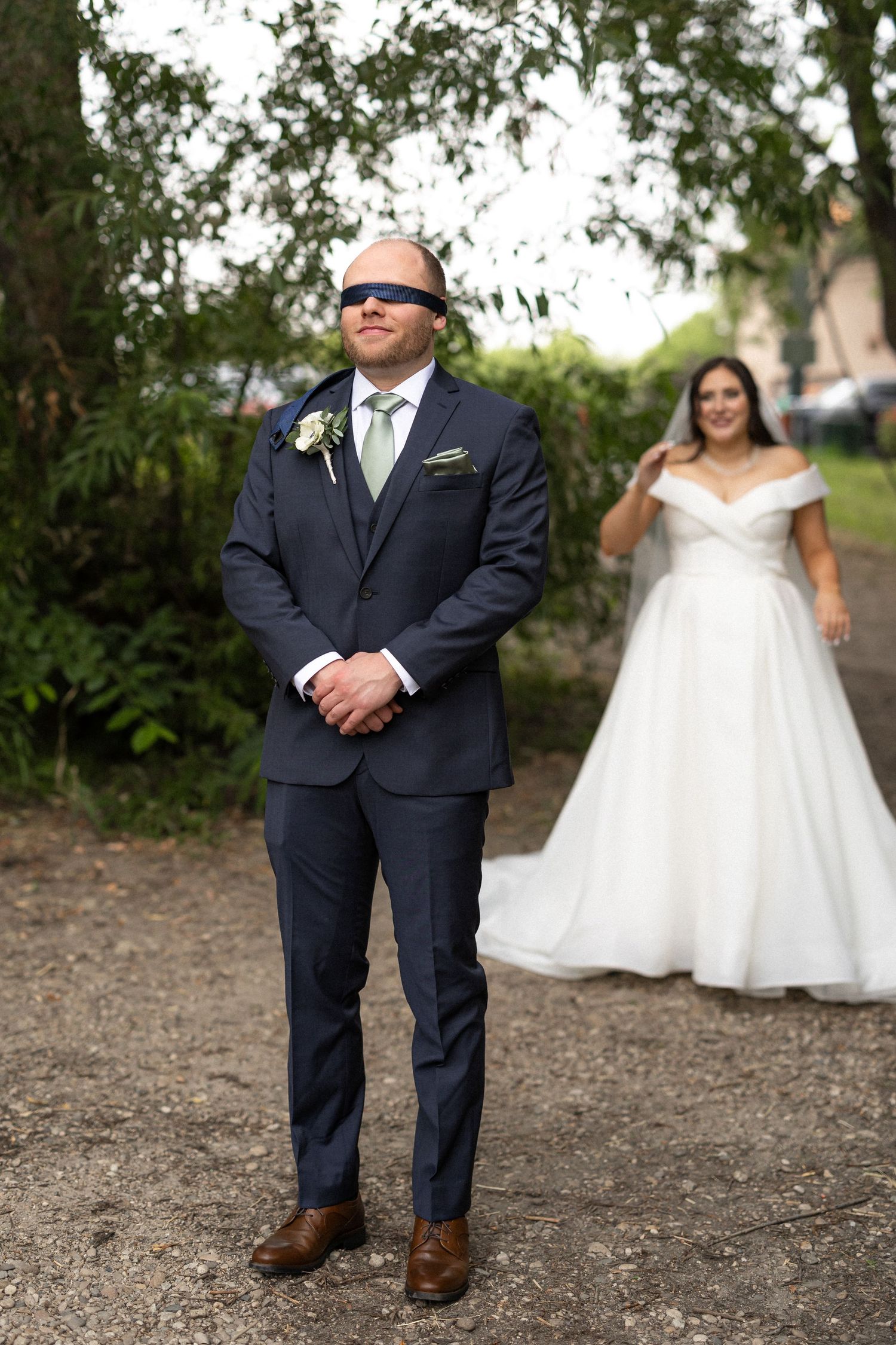 A groom in a navy suit stands on a dirt path during a wedding first look.