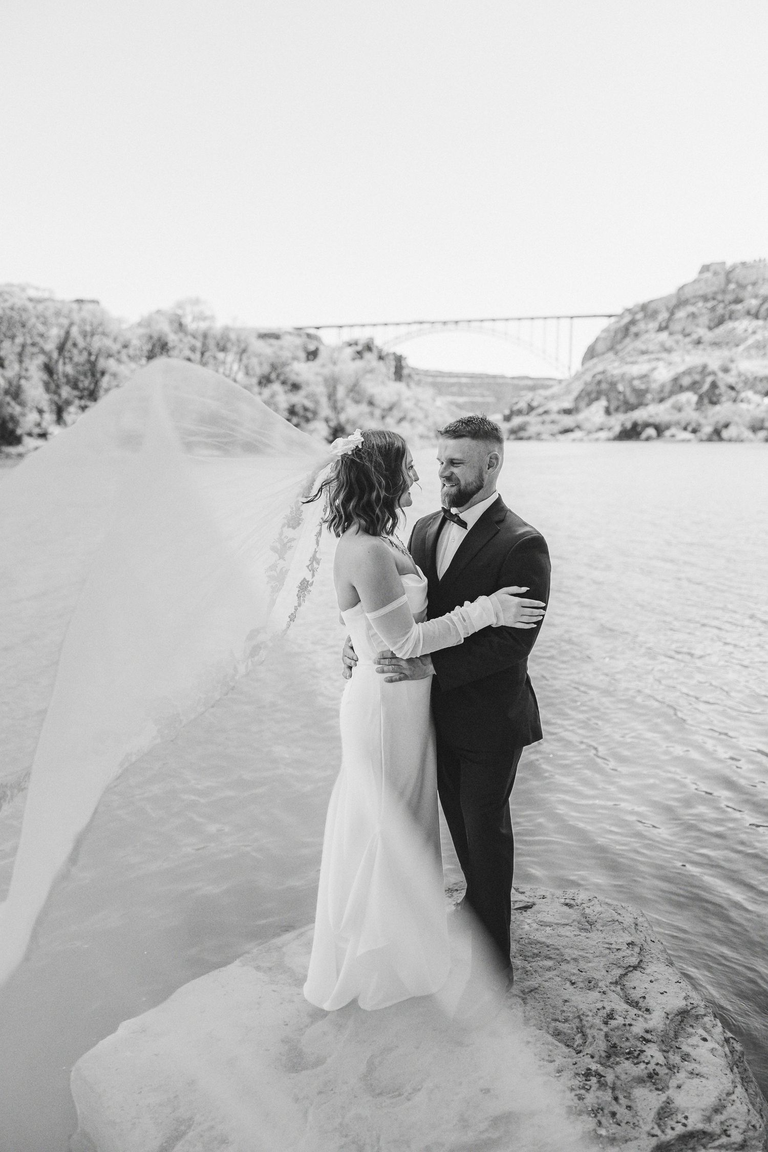 Black and white photo of bride and groom embracing by a river, veil flowing in the wind, bridge in background.