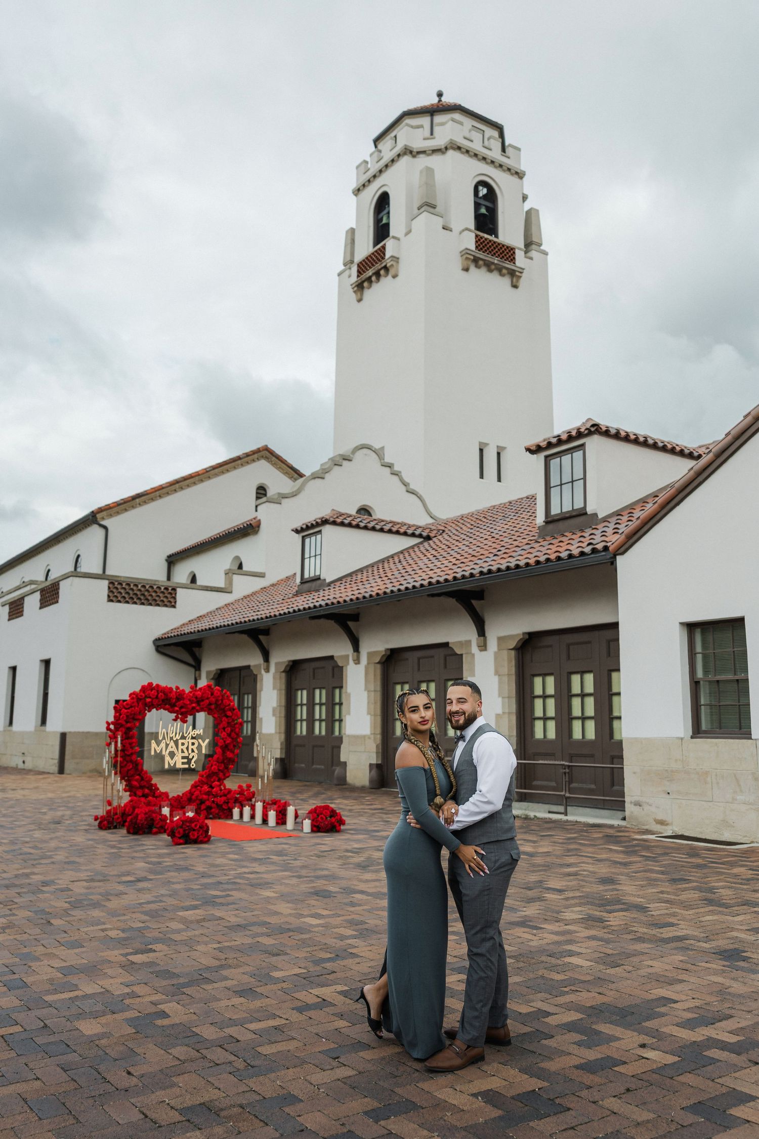Couple embraces in front of Boise Depot with a red heart-shaped floral proposal display reading Will You Marry Me.