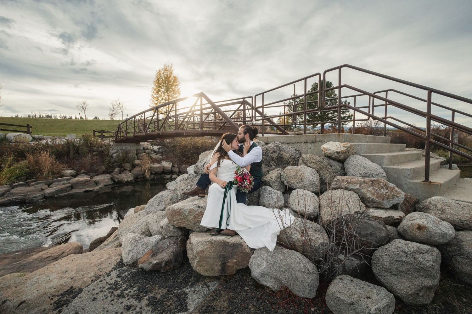 Wedding couple poses on rocky landscape with metal bridge and dramatic sky.