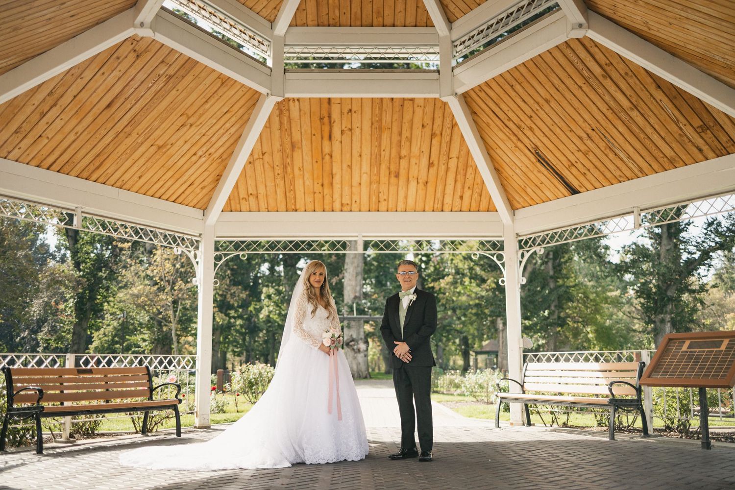 Newlyweds standing in white wooden gazebo surrounded by park benches.
