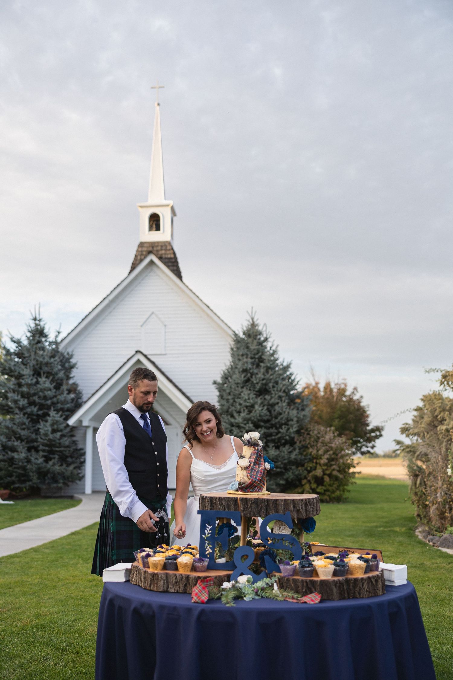 A wedding dessert table with cupcakes on wooden stands in front of a white chapel.
