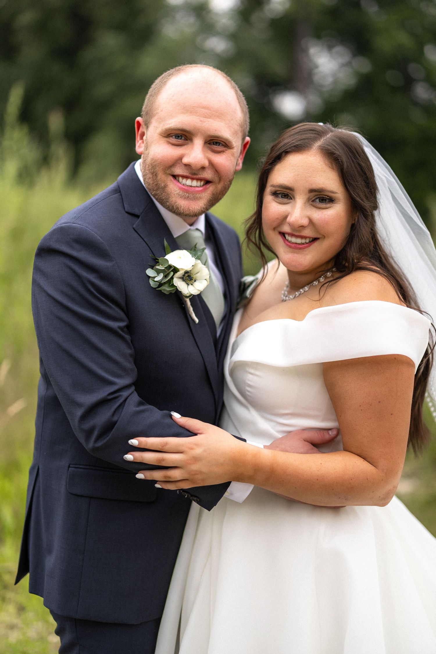 Joyful wedding couple poses outdoors in formal attire against natural green background.