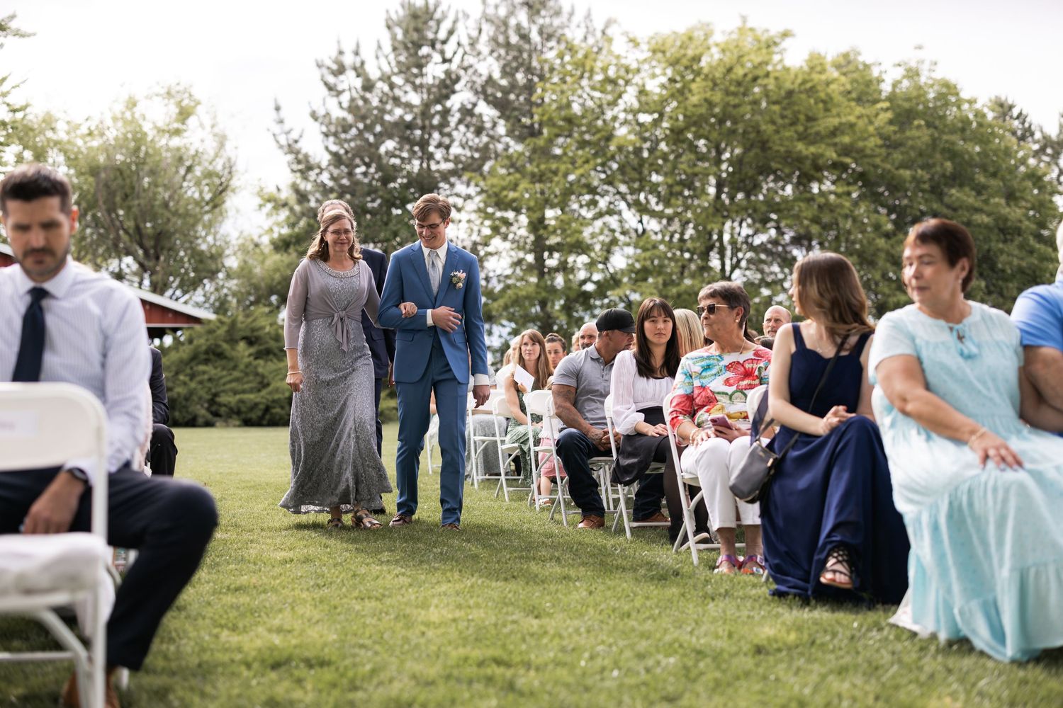 Wedding guests seated in white chairs on a lawn with trees and a red barn in the background.