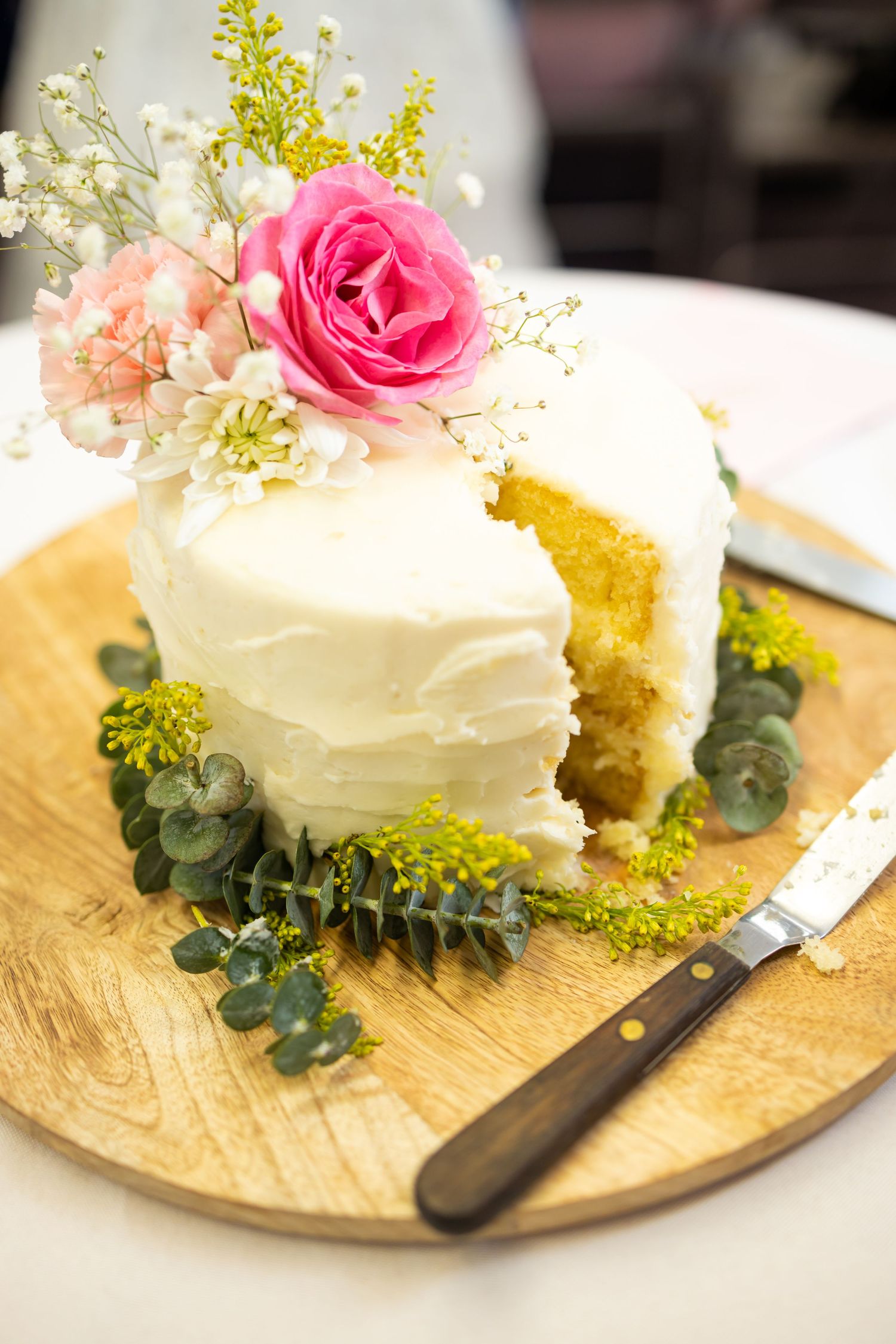 A white wedding cake slice decorated with pink roses and eucalyptus on a wooden serving board.