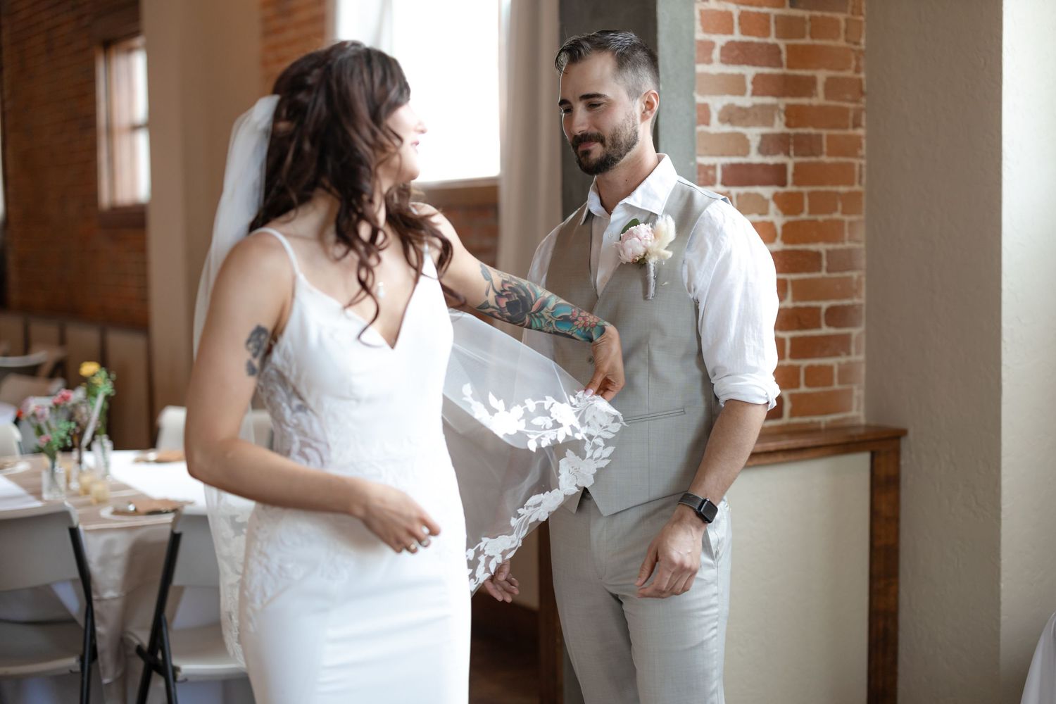 Tender moment between wedding couple in front of rustic brick wall with natural light streaming in.