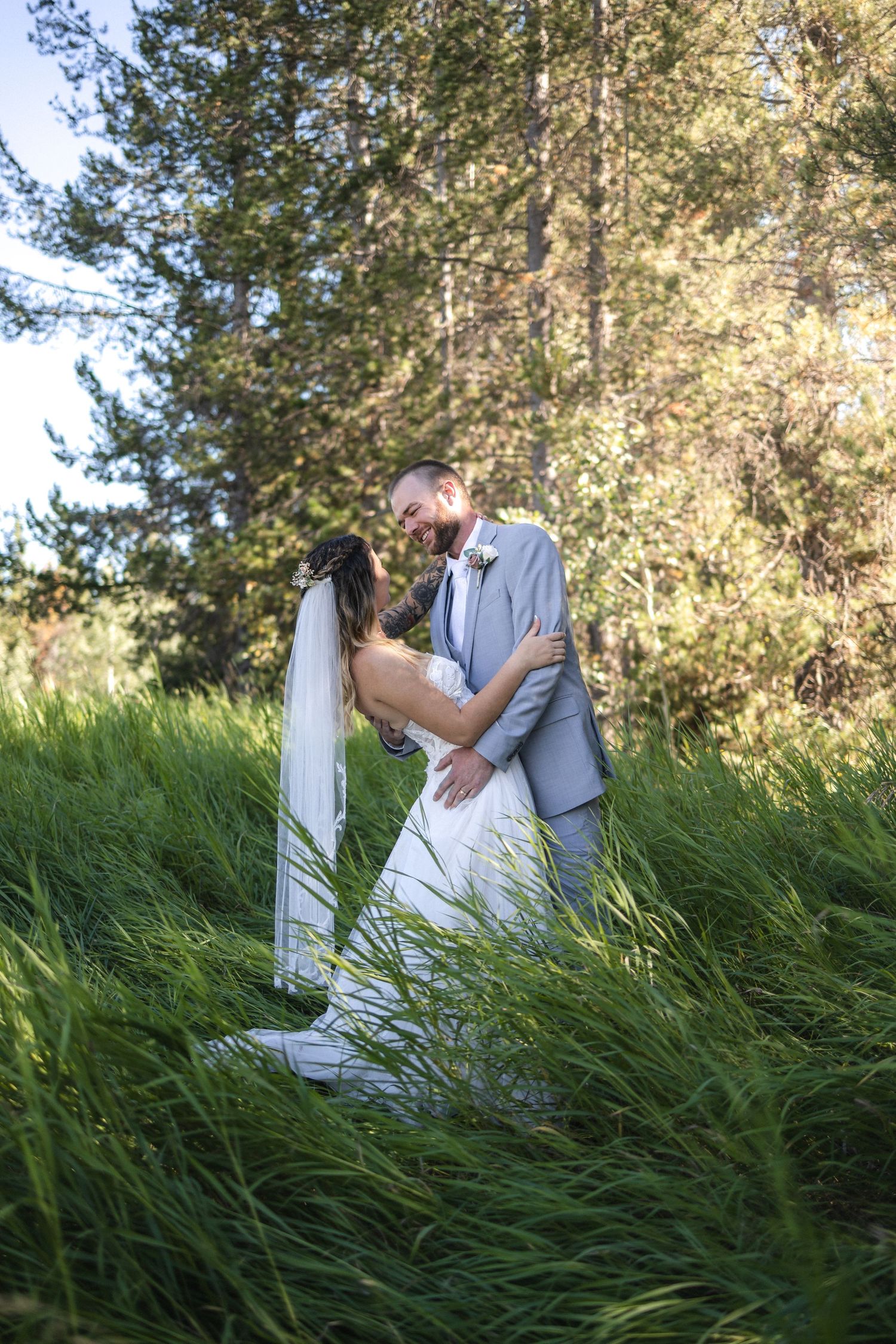A romantic wedding moment captured in tall grass with sunlit forest in the background.
