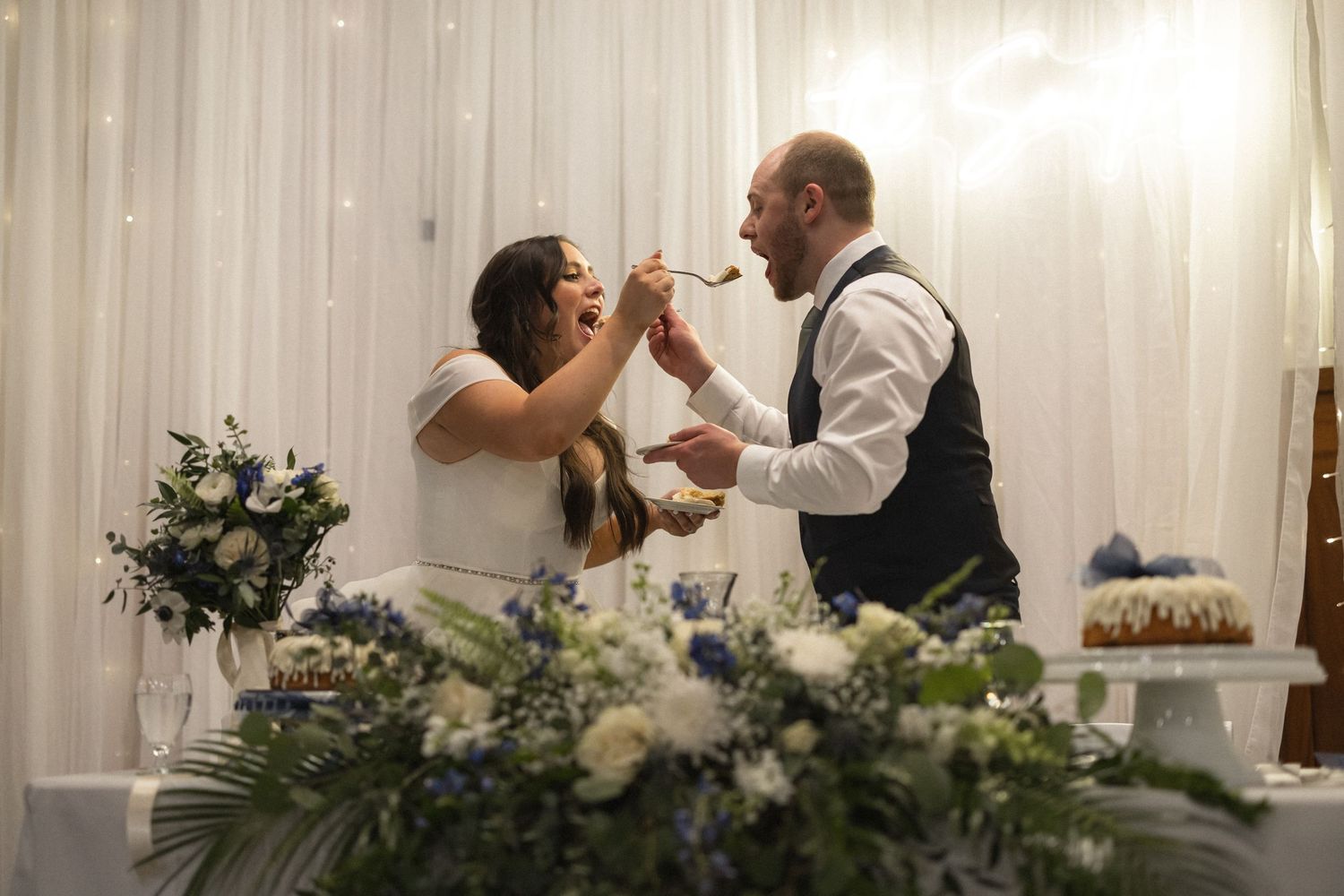Playful moment during wedding reception as couple shares dessert at table with floral decorations.