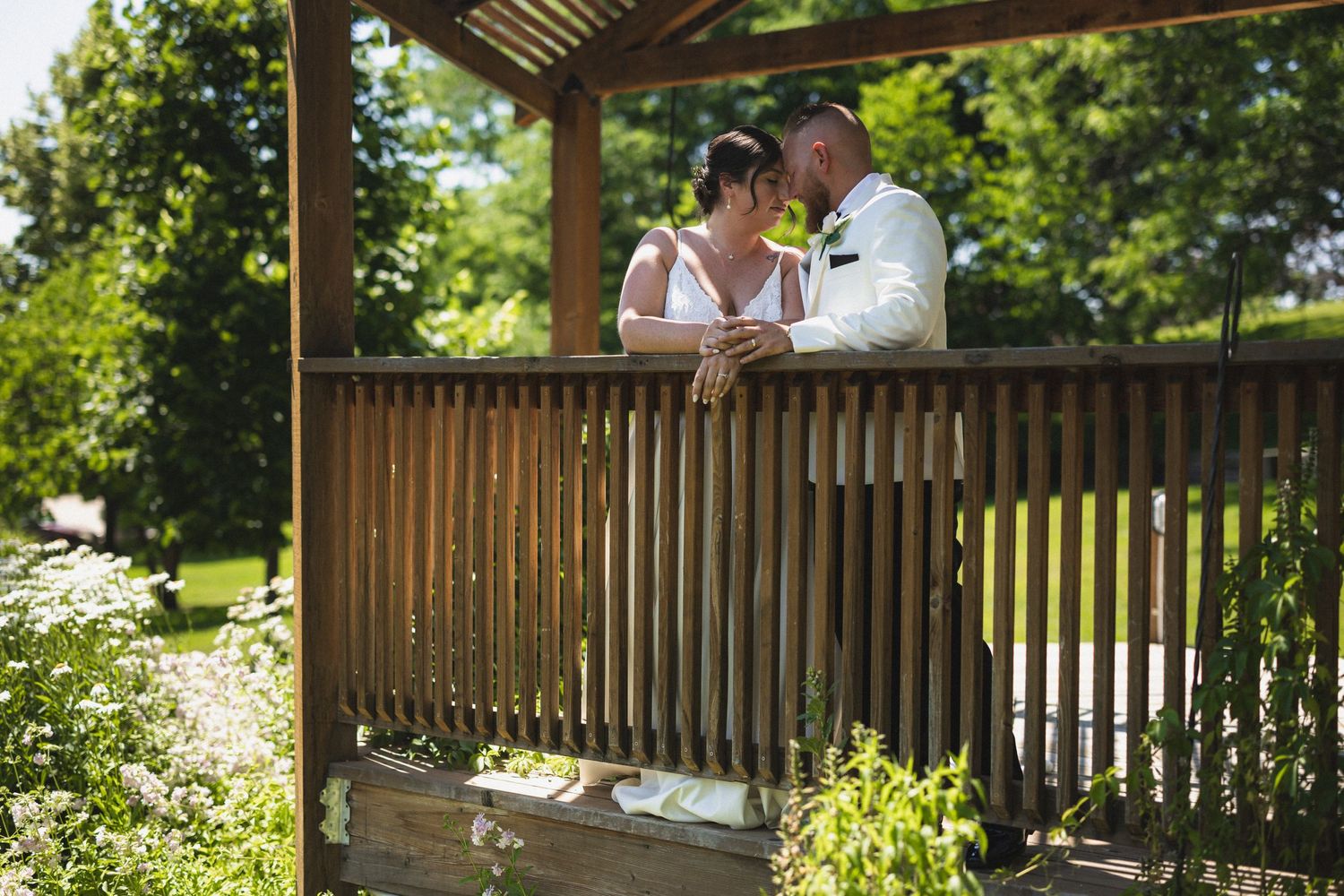 Wedding couple shares an intimate moment under a wooden pergola surrounded by greenery.