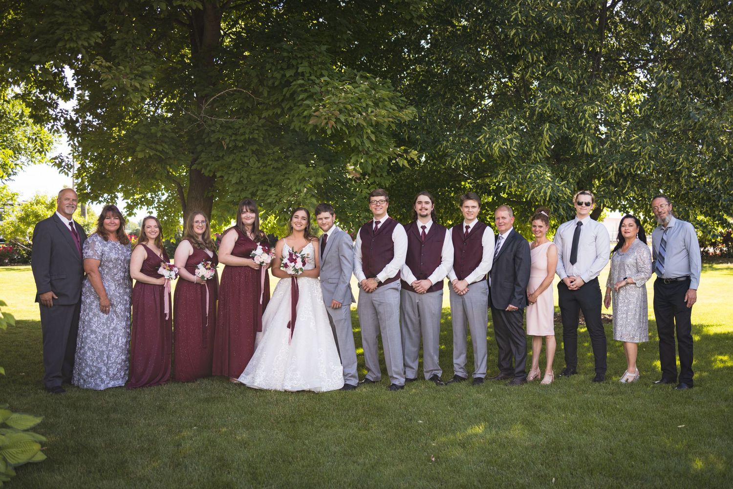 A wedding party in burgundy and gray attire poses together on a grassy lawn under large trees.