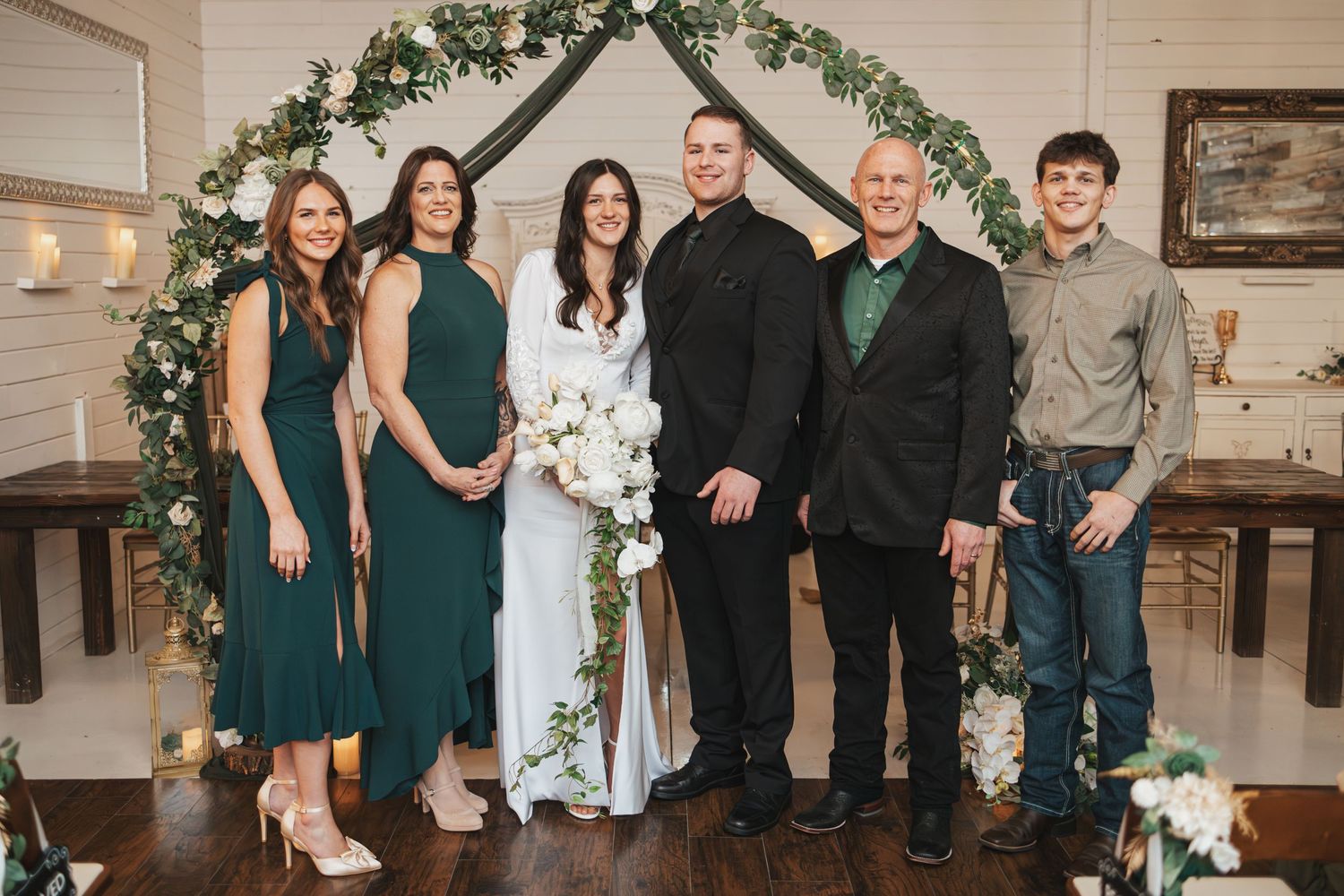 Wedding group photo under floral arch with emerald green and black formal attire.