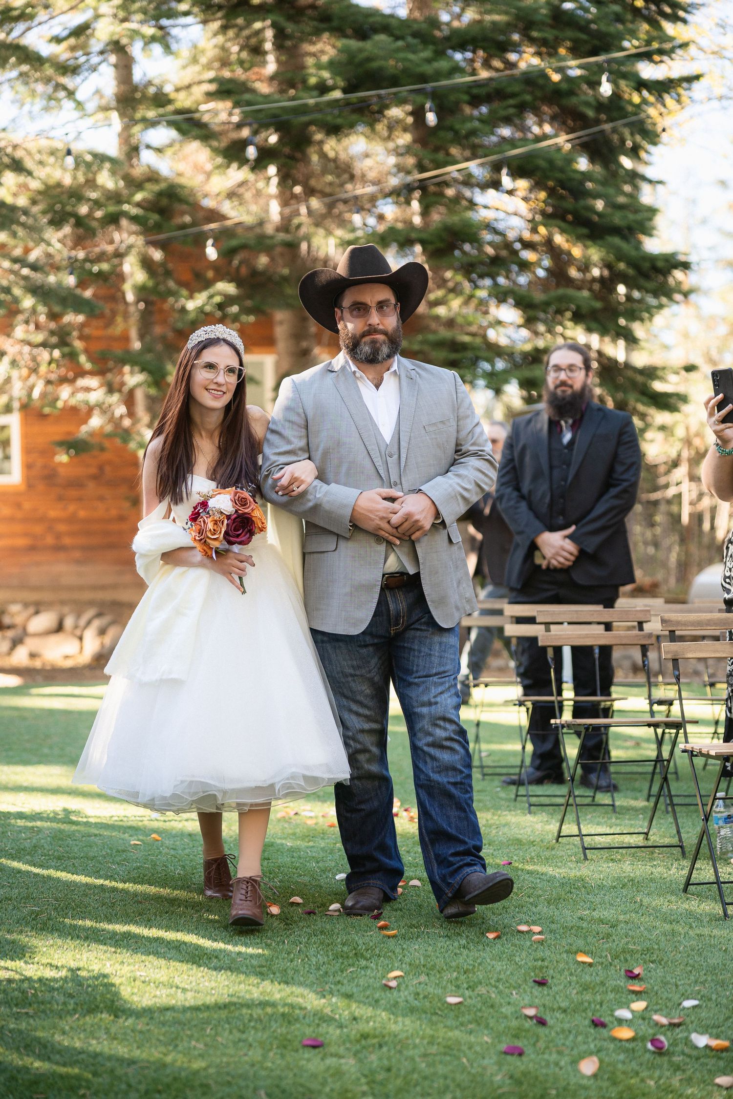 Western-themed wedding processional with groomsman in cowboy hat escorting bride across lawn.