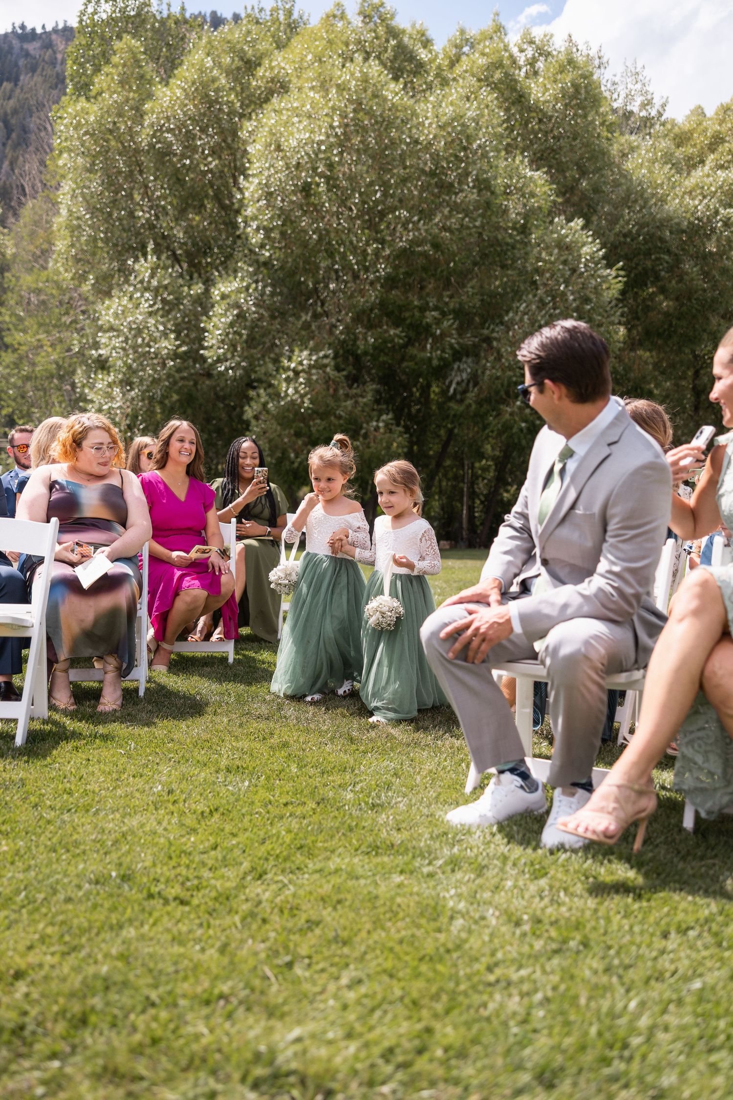 Wedding guests seated on grass enjoying outdoor celebration with trees in background.