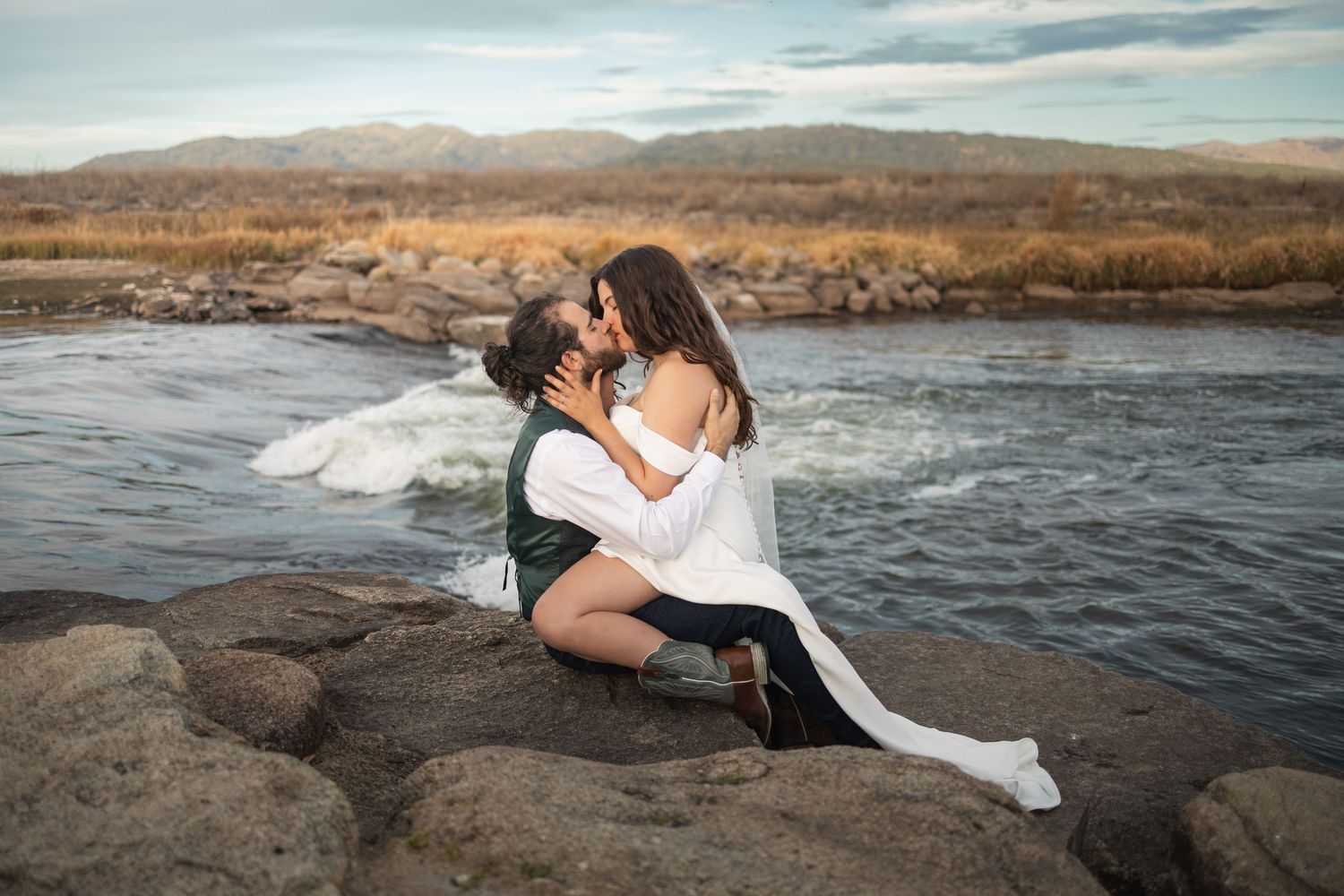 Couple in white embraces on rocky riverbank with mountains and flowing water in scenic natural landscape.