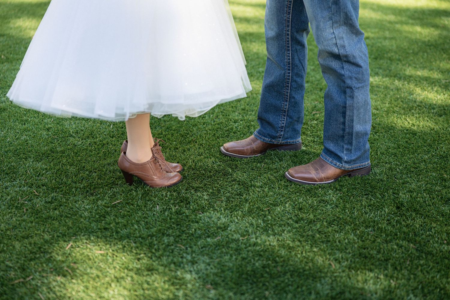 Wedding couple's feet in western boots with bride wearing short tulle dress on green grass.