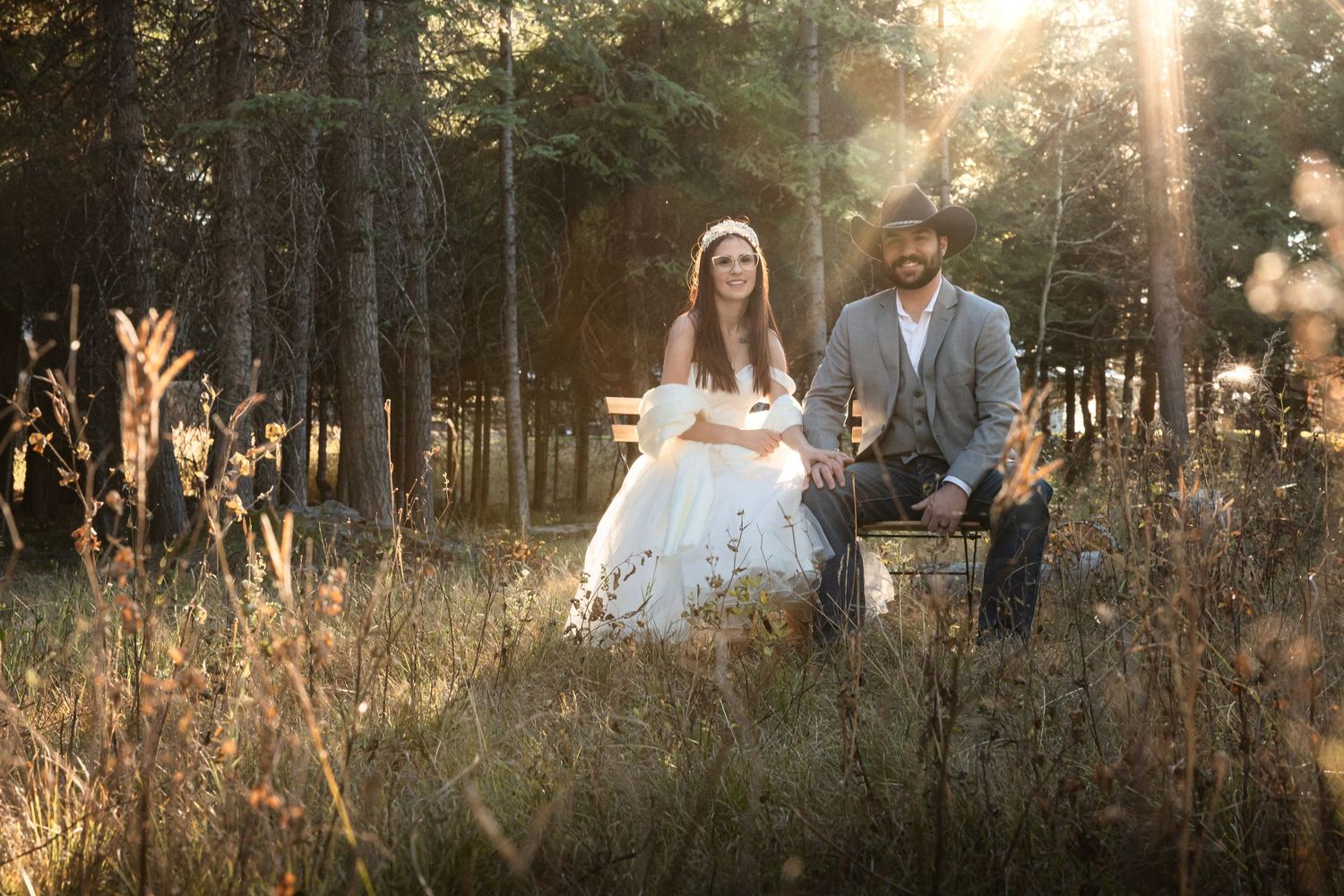 Wedding couple sits together in tall grass during sunset in a pine forest.