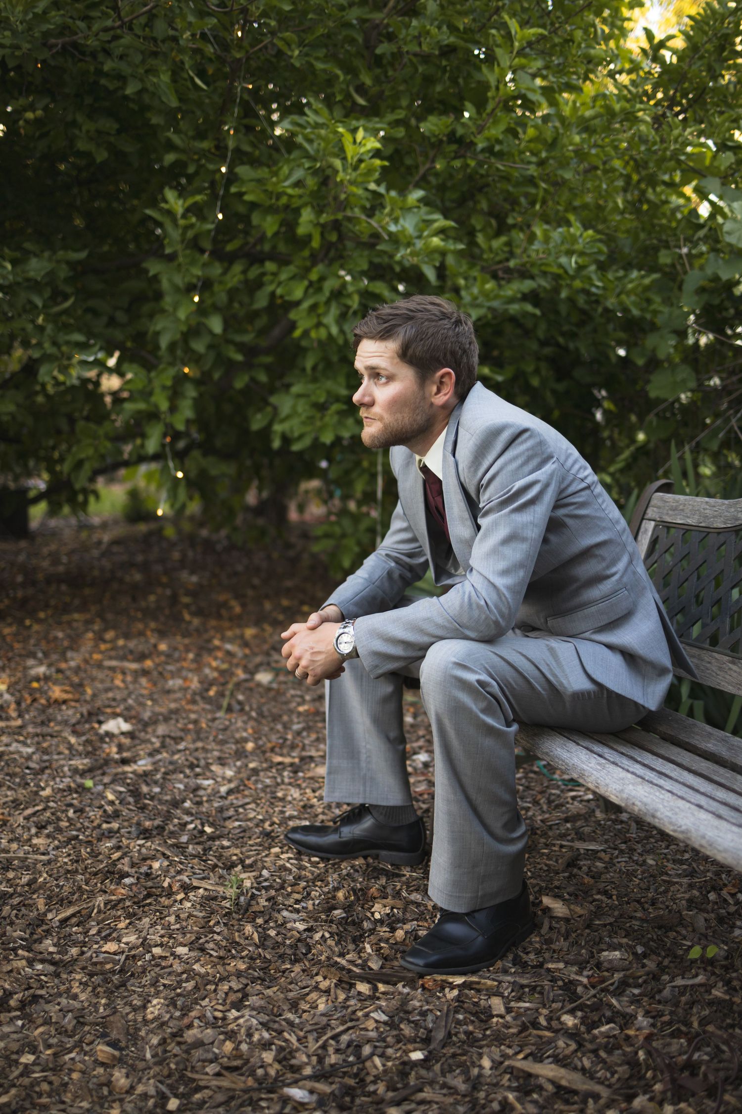 A person in a grey suit sits contemplatively on a wooden bench in a garden setting.
