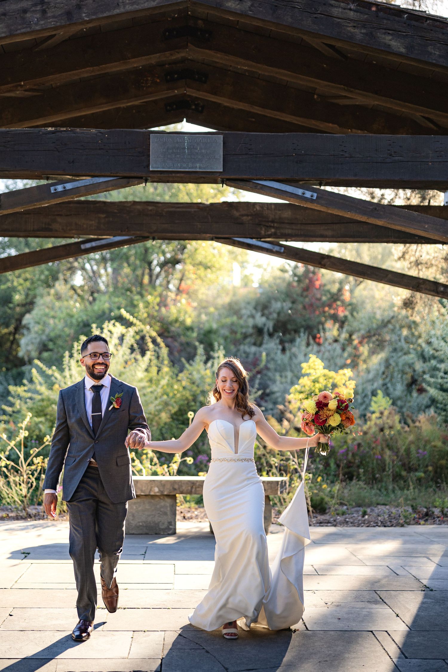 Joyful newlyweds walk hand in hand beneath wooden pergola after outdoor wedding ceremony.