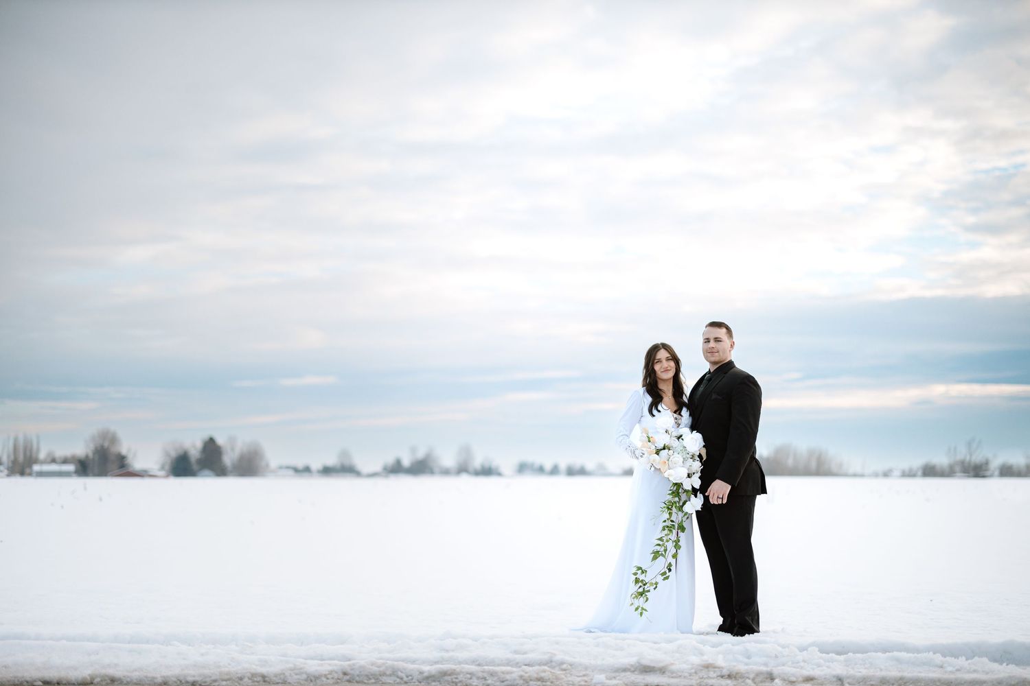 Wedding couple embraces on snowy field with overcast winter sky in background.