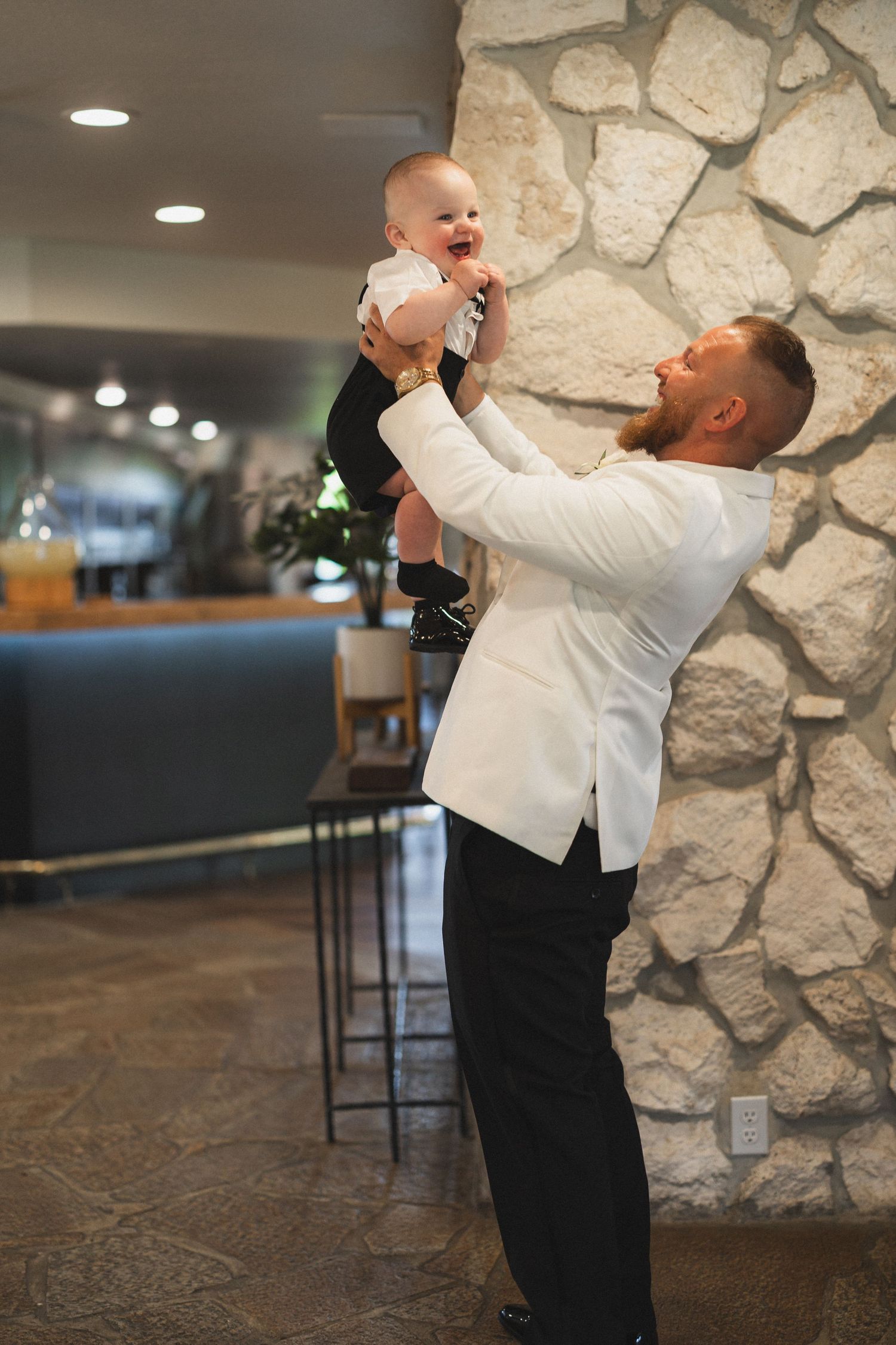 A person in formal wear lifting a baby high in the air against a stone wall backdrop.