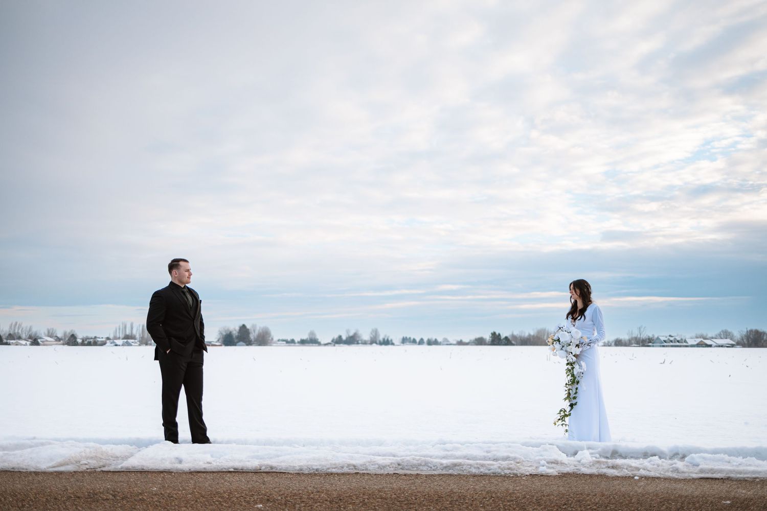 Couple stands apart in snowy winter landscape during wedding photoshoot.