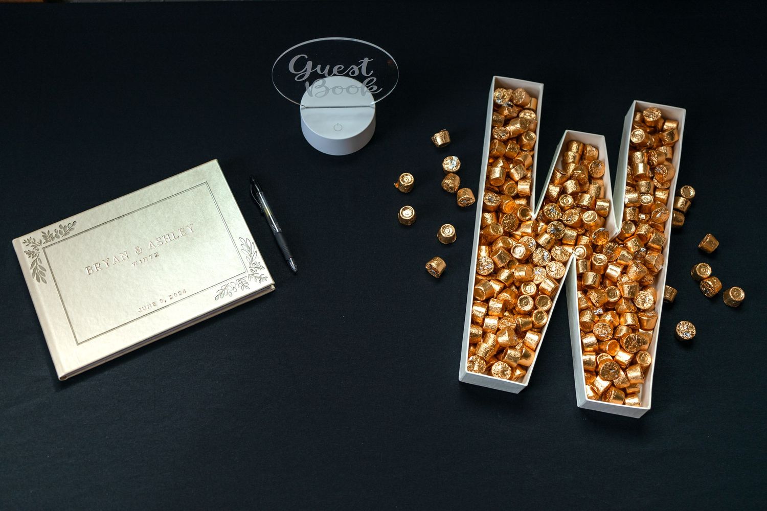 Letter W made of gold sequins with notecard and drink on dark background.