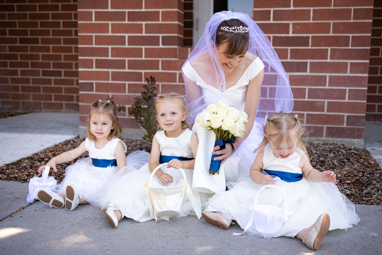Bride with flower girls in white dresses with blue sashes sitting on brick steps.